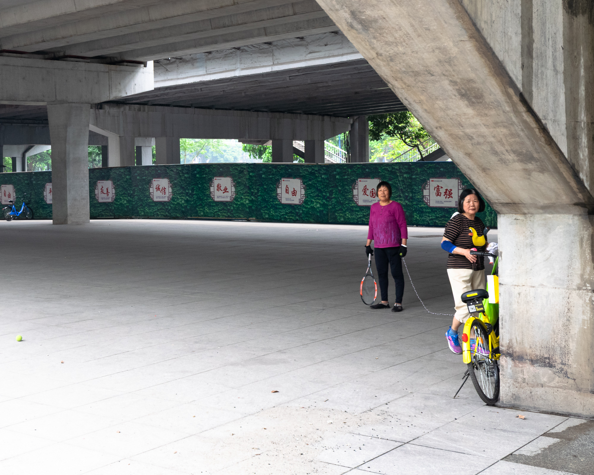 Two women with a tennis racket and shared bike under an overpass, Guangzhou China