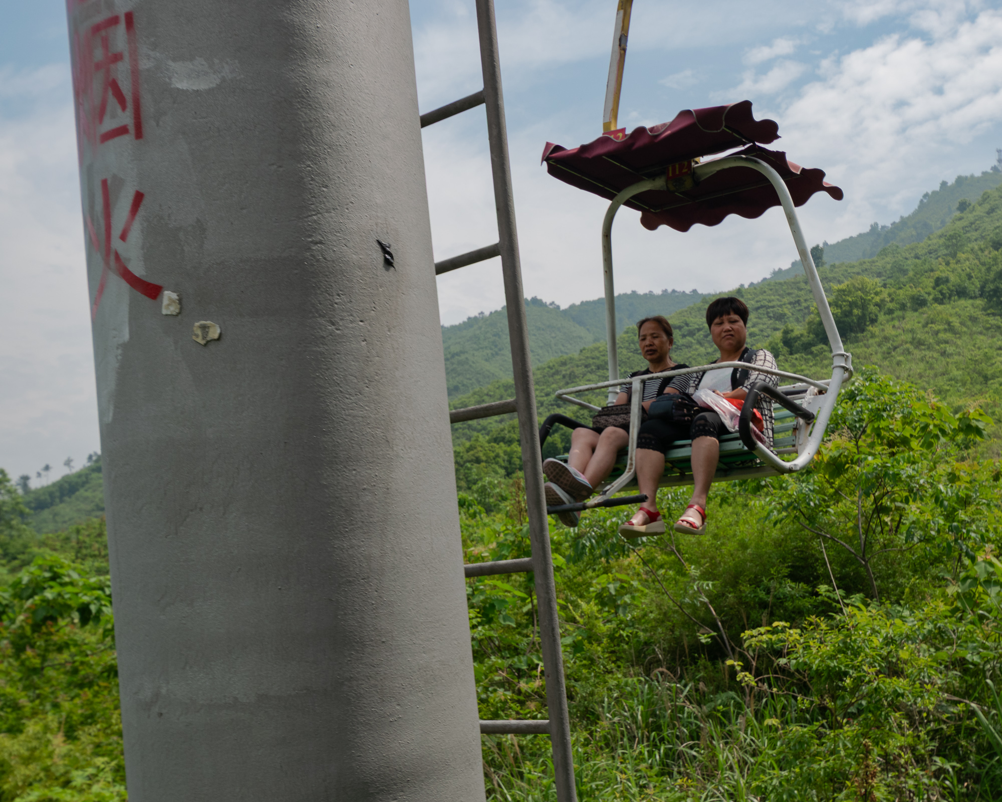 Two women ride a chairlift above forested hills in China — pastoral, elevated view