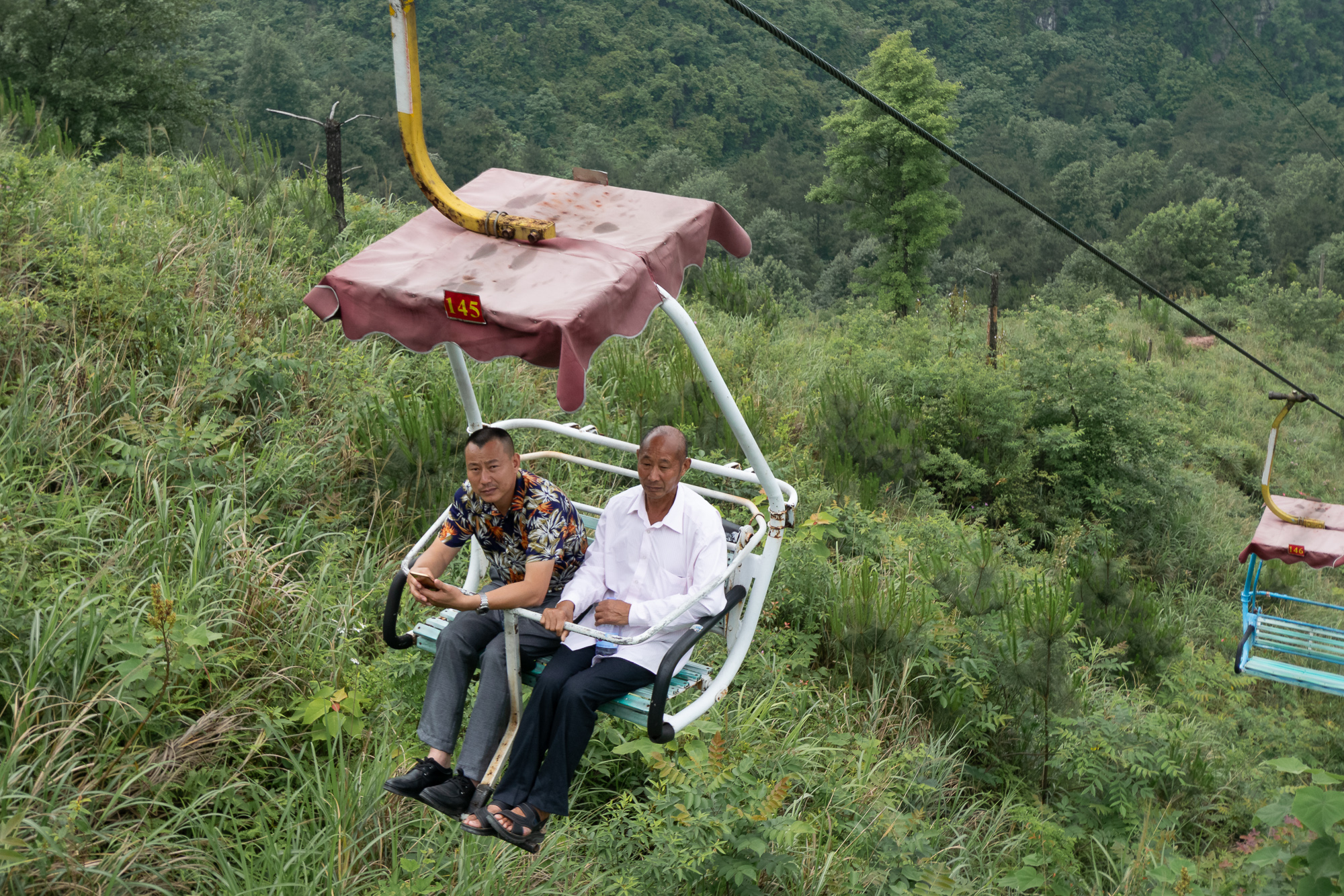 Two men ride a worn chairlift numbered 145 above tall grass and shrubs, with forested hills behind them. One wears a floral shirt and checks his phone; the other wears a white shirt and dark trousers.