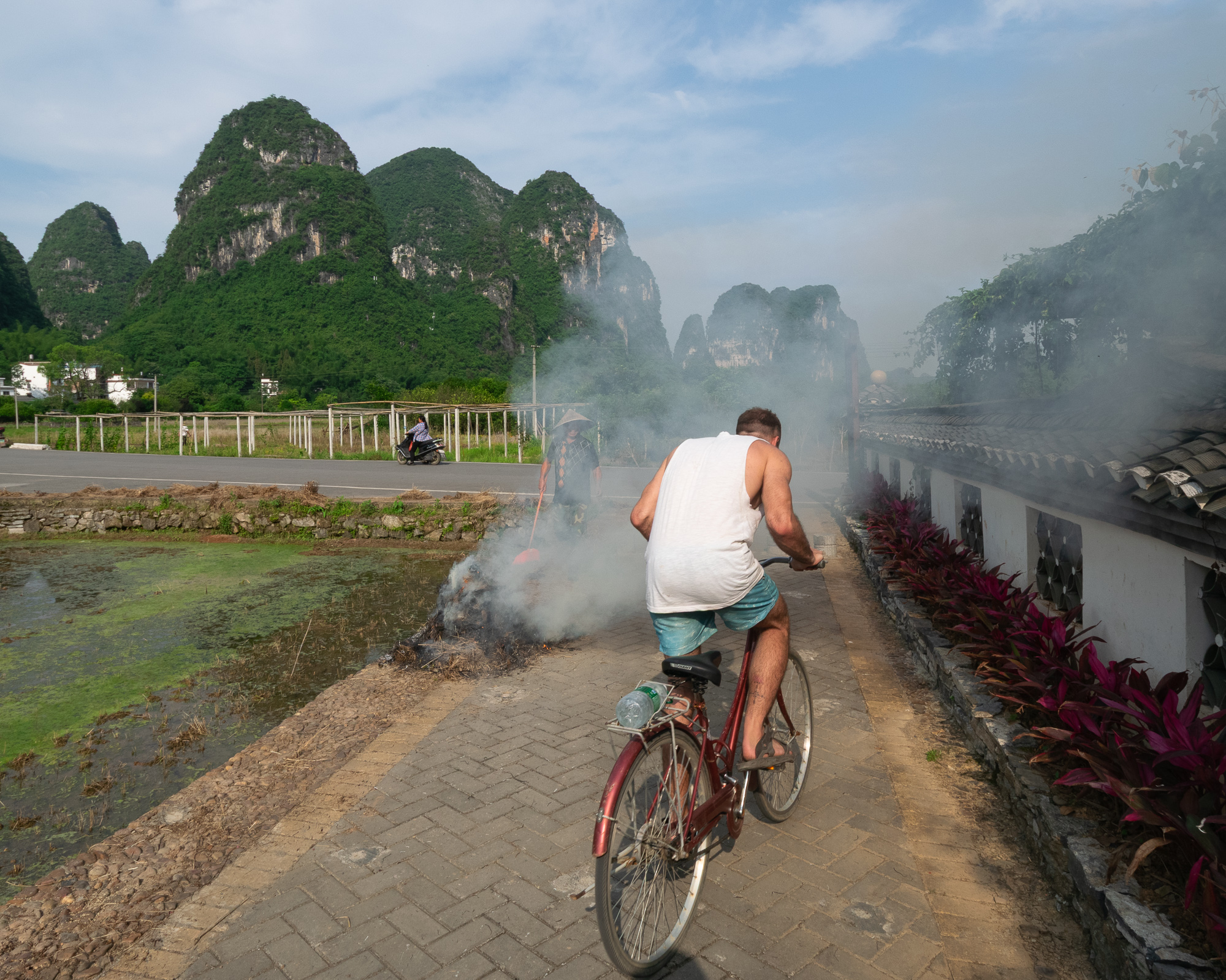 A man in a white tank top and blue shorts rides a red bicycle along a brick path past a smoldering pile of brush, while a woman in a conical hat tends the fire and karst mountains rise in the background.