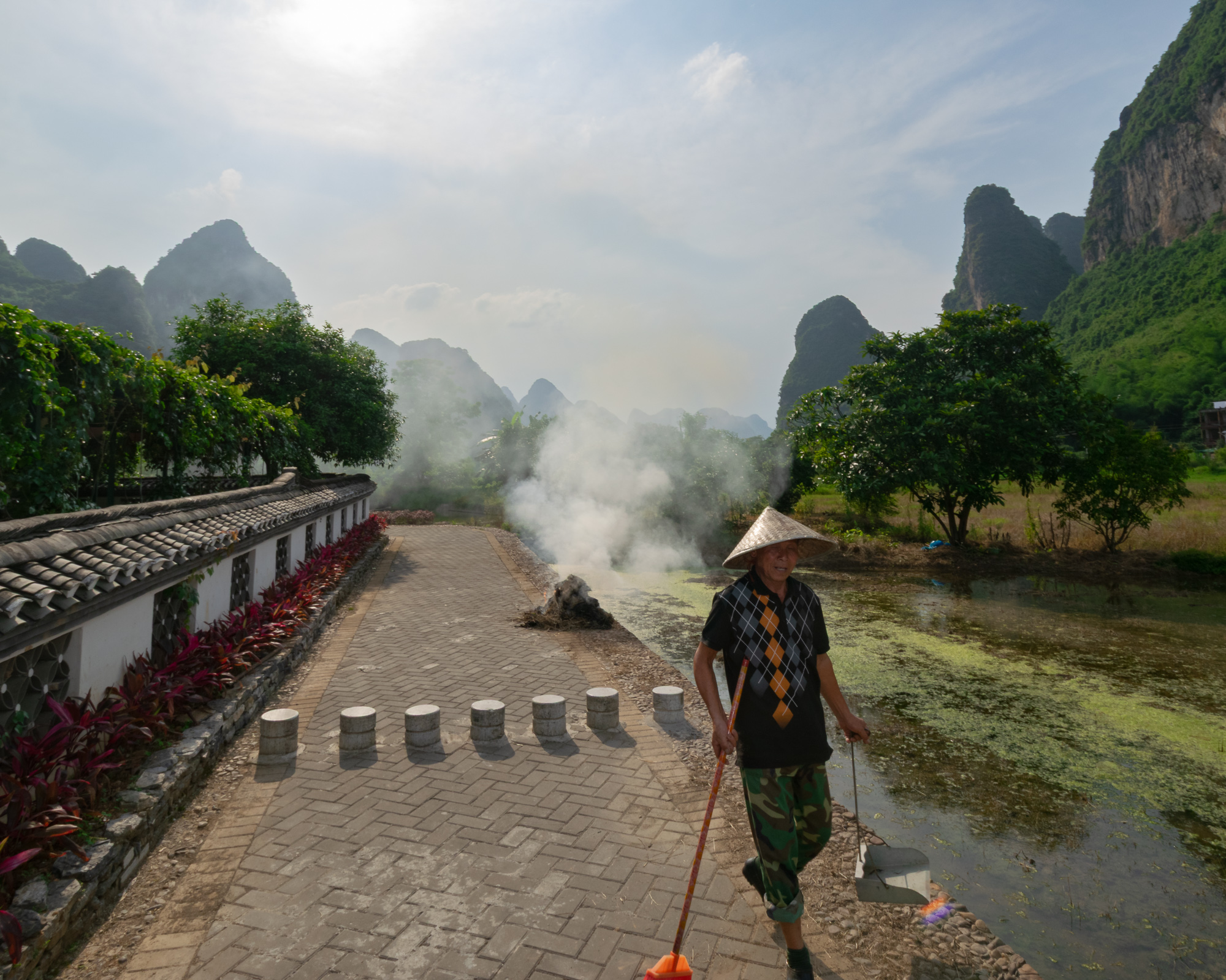 A worker in a conical straw hat and camouflage trousers walks along a brick path beside a smoking pile of brush, with karst mountain peaks rising in the background.