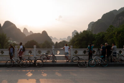 A group of people stand with their bicycles along a white stone bridge railing at dusk, looking out at the misty karst mountain peaks of Yangshuo, China. The hazy sun lowers behind the jagged silhouettes while cyclists pause to take in the view.