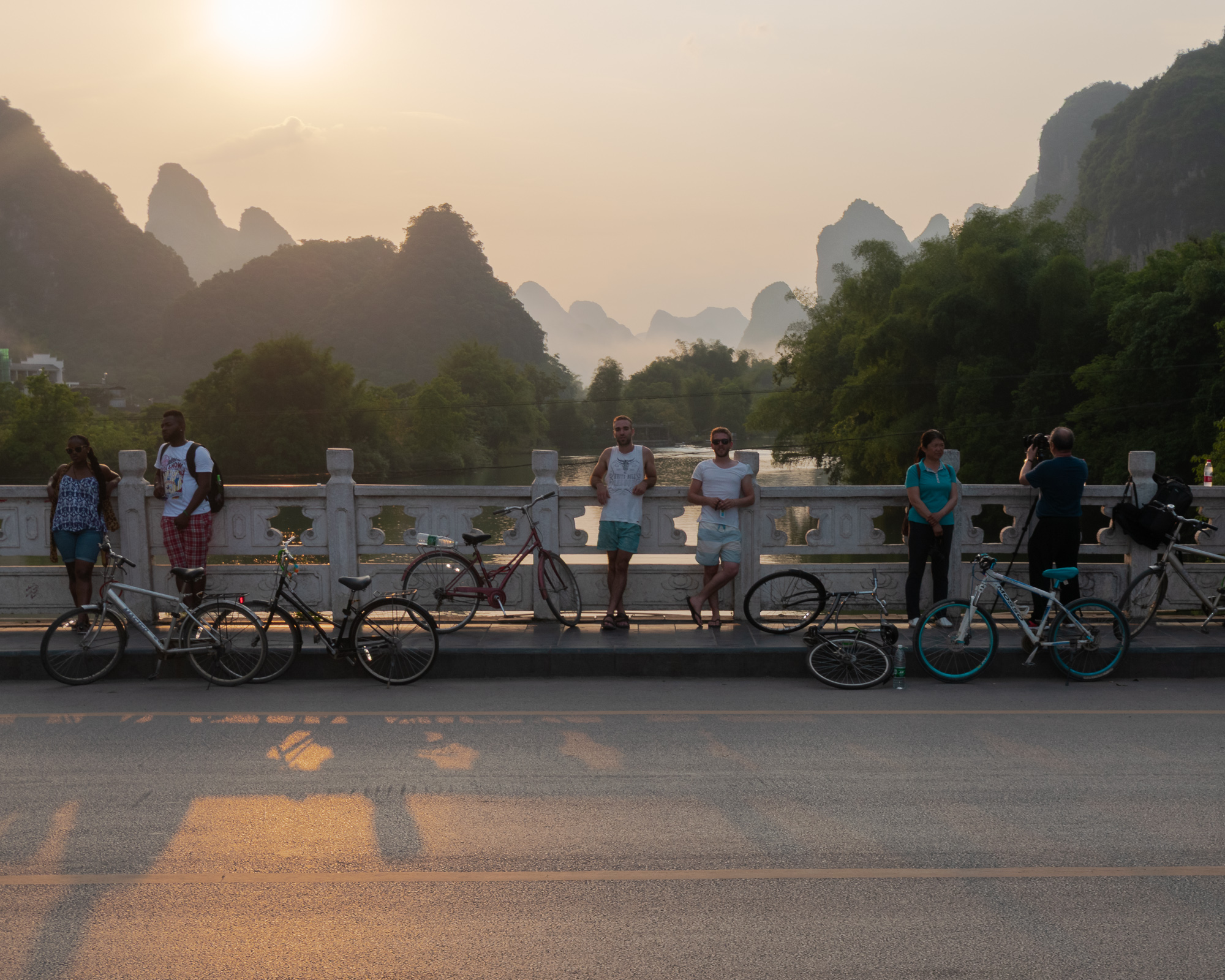 Travelers and locals lean against the stone balustrade of a bridge with parked bicycles at their feet, as karst mountain peaks rise through hazy light above a river at sunset.