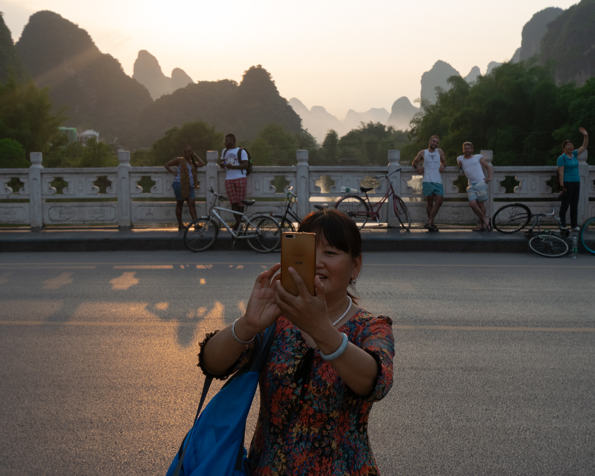A woman in a floral dress holds up a gold smartphone to take a photo on a bridge at sunset, while tourists and parked bicycles line the stone railing behind her with karst mountain peaks rising in the hazy distance.