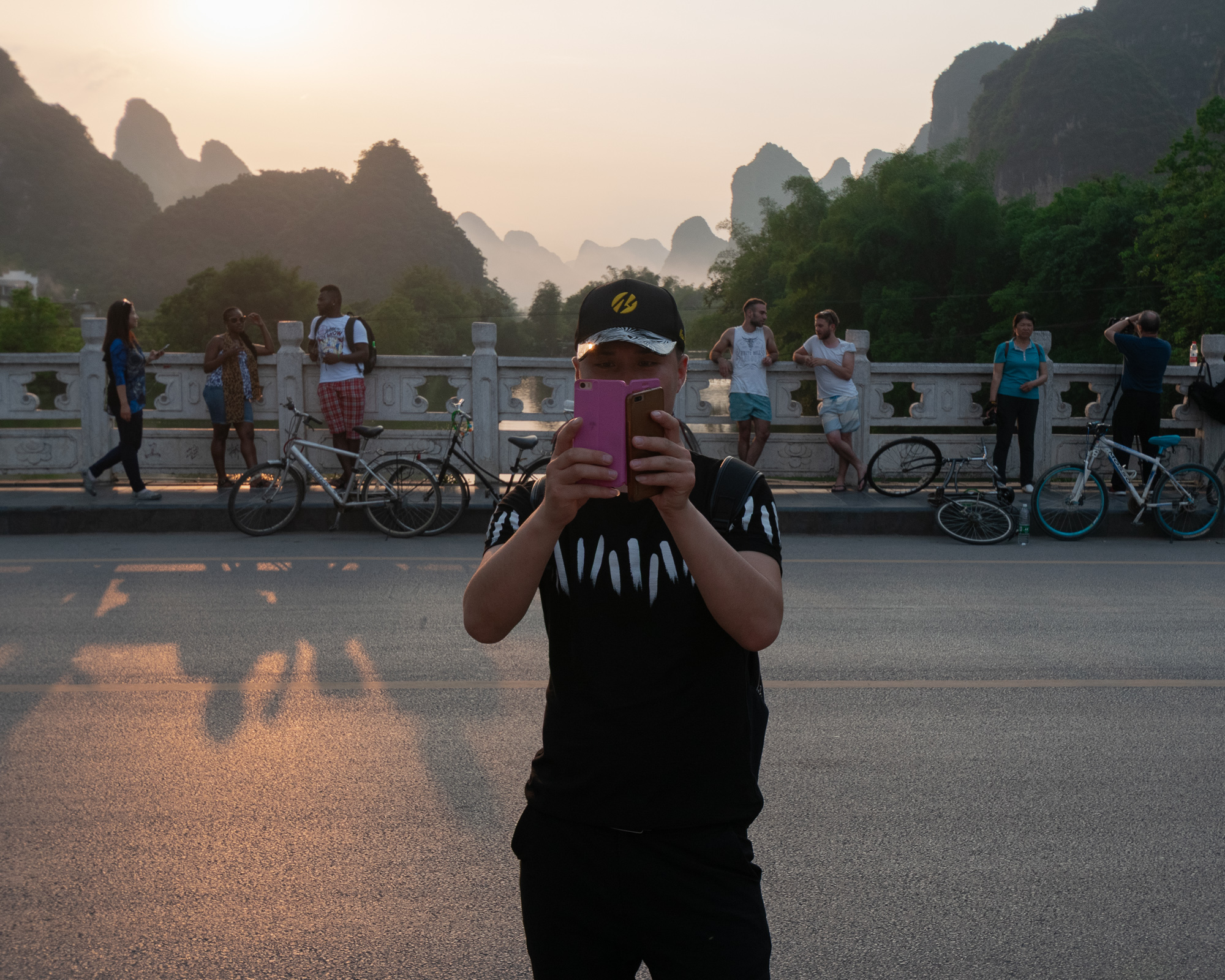 A man in a black cap and shirt holds two smartphones to his face on a bridge, photographing the viewer. Behind him, tourists lean on a stone balustrade with bicycles, karst peaks rising in hazy sunset light.