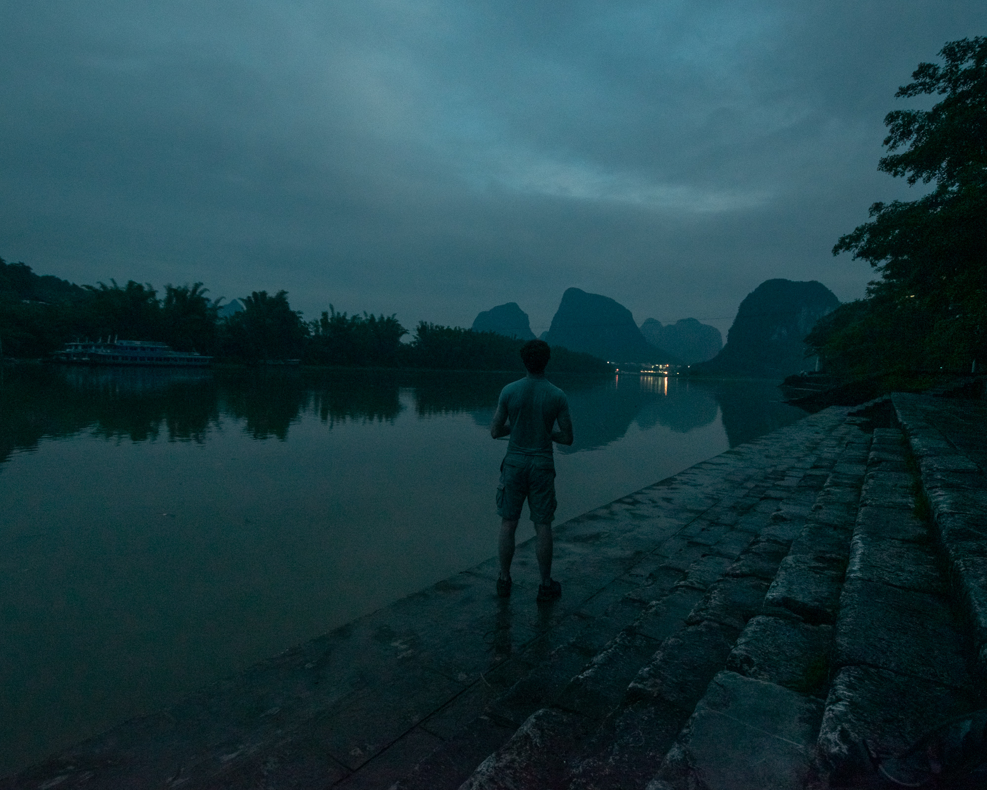 A man in shorts and a t-shirt stands on a wet stone embankment beside a still river at dusk, facing distant karst mountains under heavy clouds. Lights from a far shore reflect on the water, and a docked boat sits along the opposite bank.