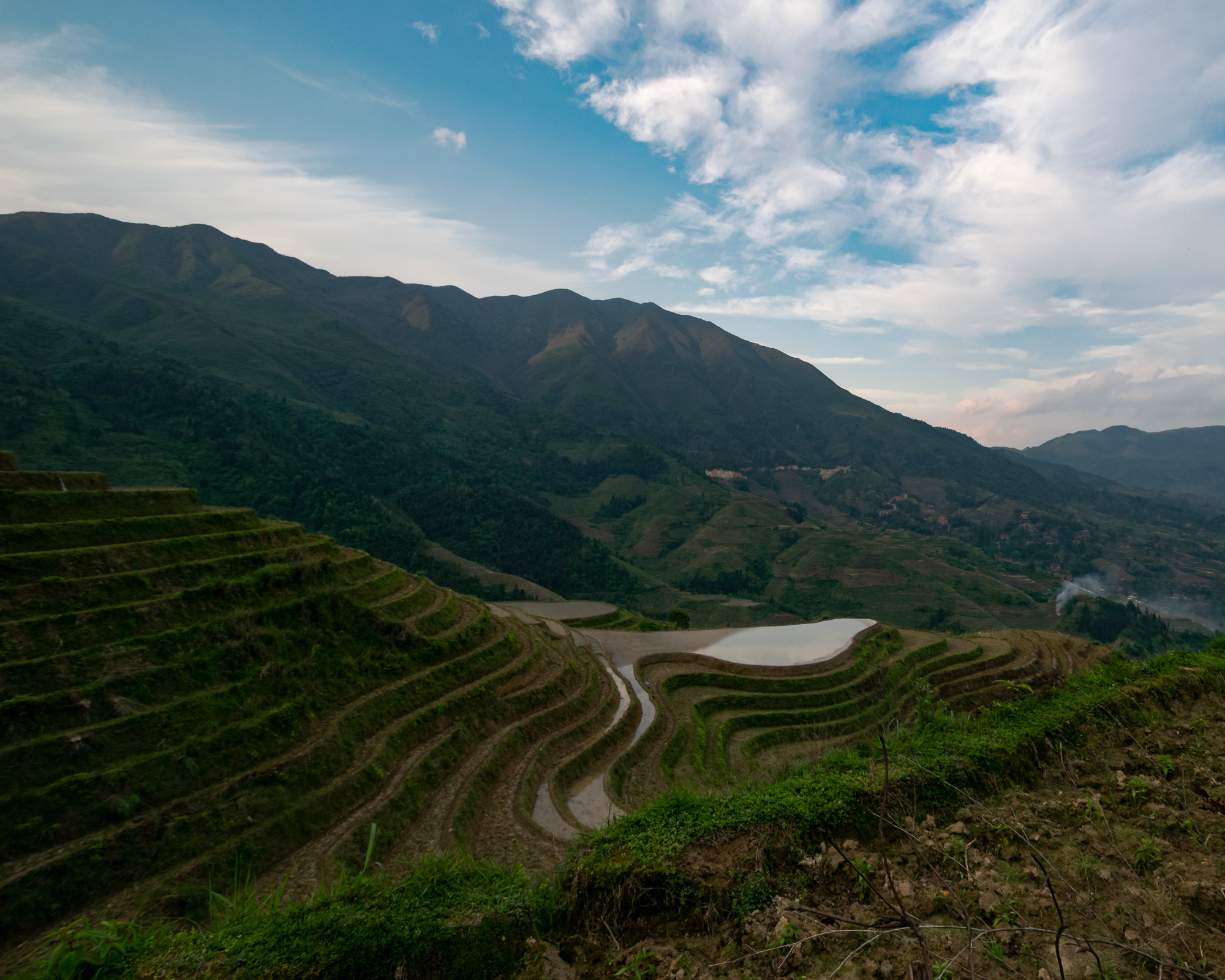 Curved, flooded rice terraces descend a hillside in Guangxi, China, with forested mountains and a partly cloudy sky behind them. A distant village clings to the slope on the right side of the valley.