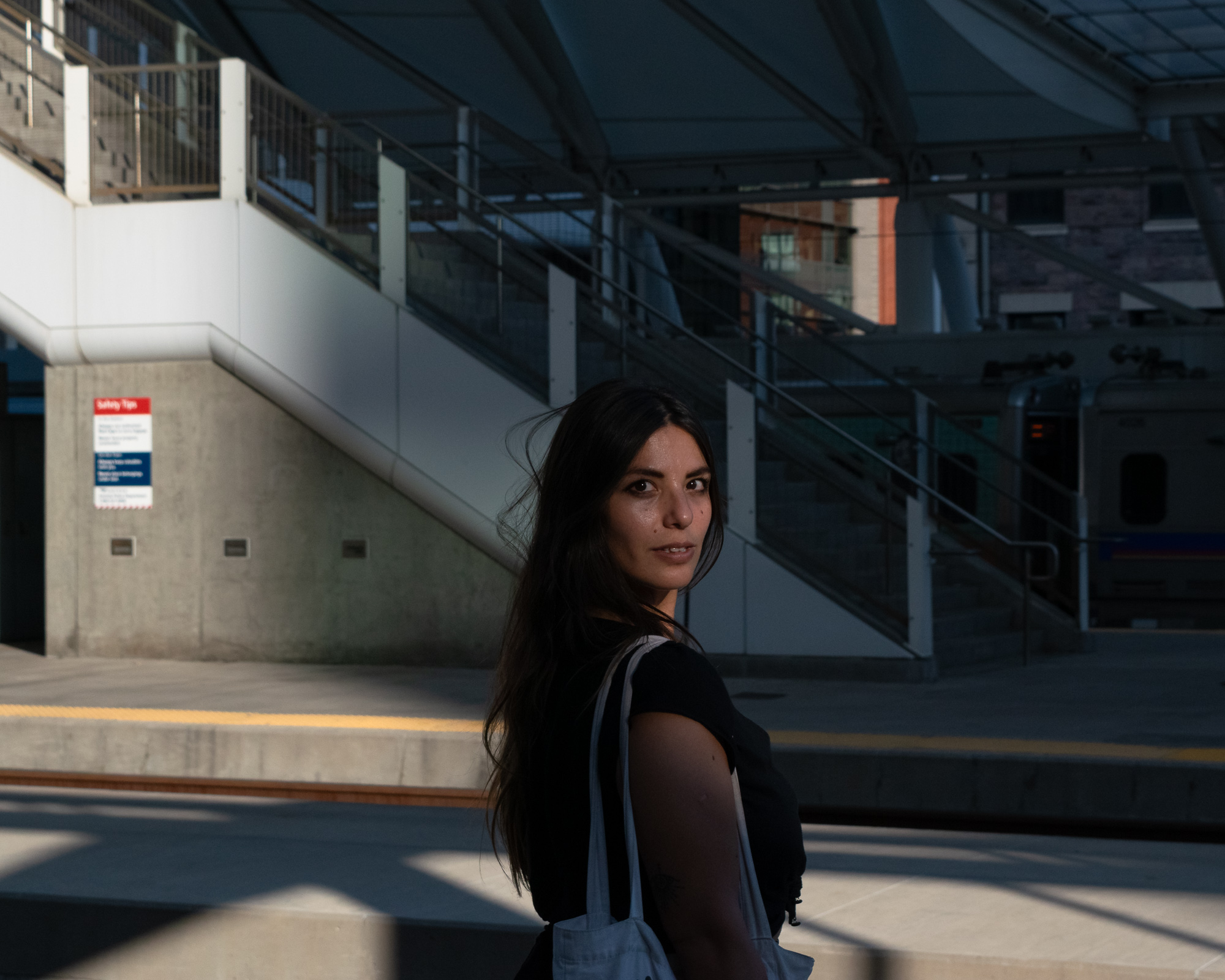 A woman at a Denver transit platform, caught in dramatic directional light, looking back at the camera — digital