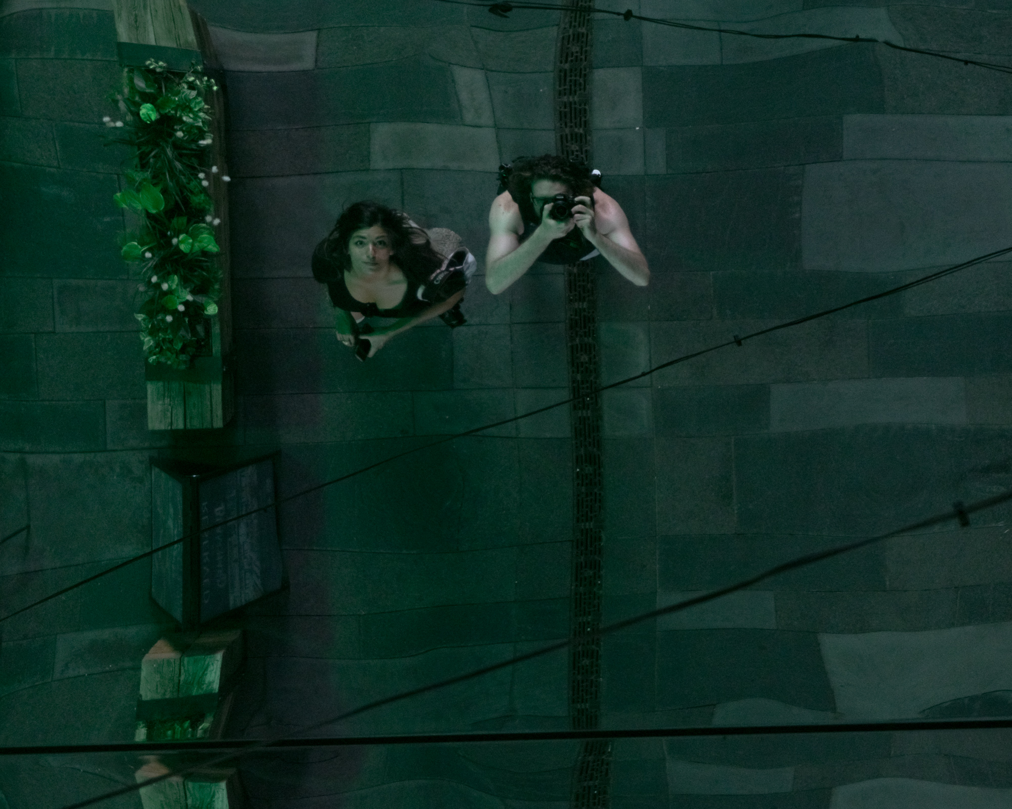 Looking straight down at a reflective rooftop in Denver — two figures seen from directly above, wires crossing, deep green tones, disorienting perspective — digital