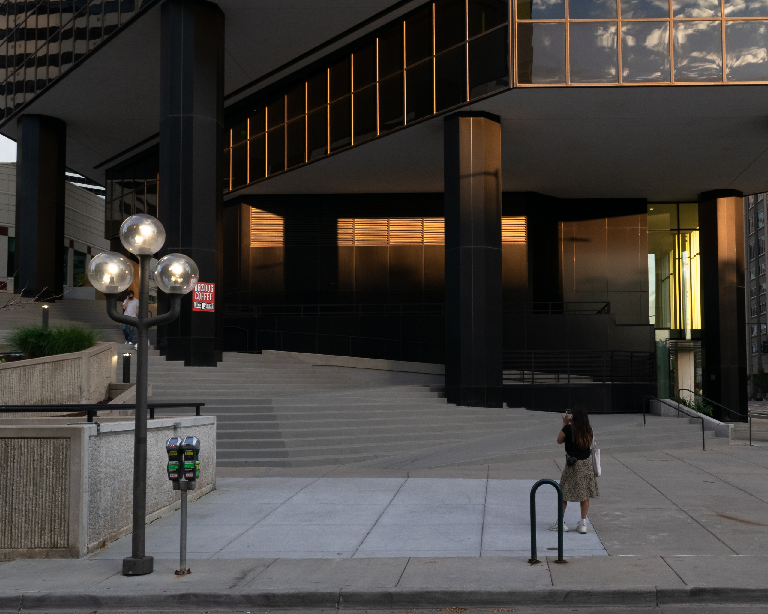 A city plaza at dusk in Denver — dark brutalist columns, warm amber light glowing from a modernist building interior, a lone figure standing at the base of the steps