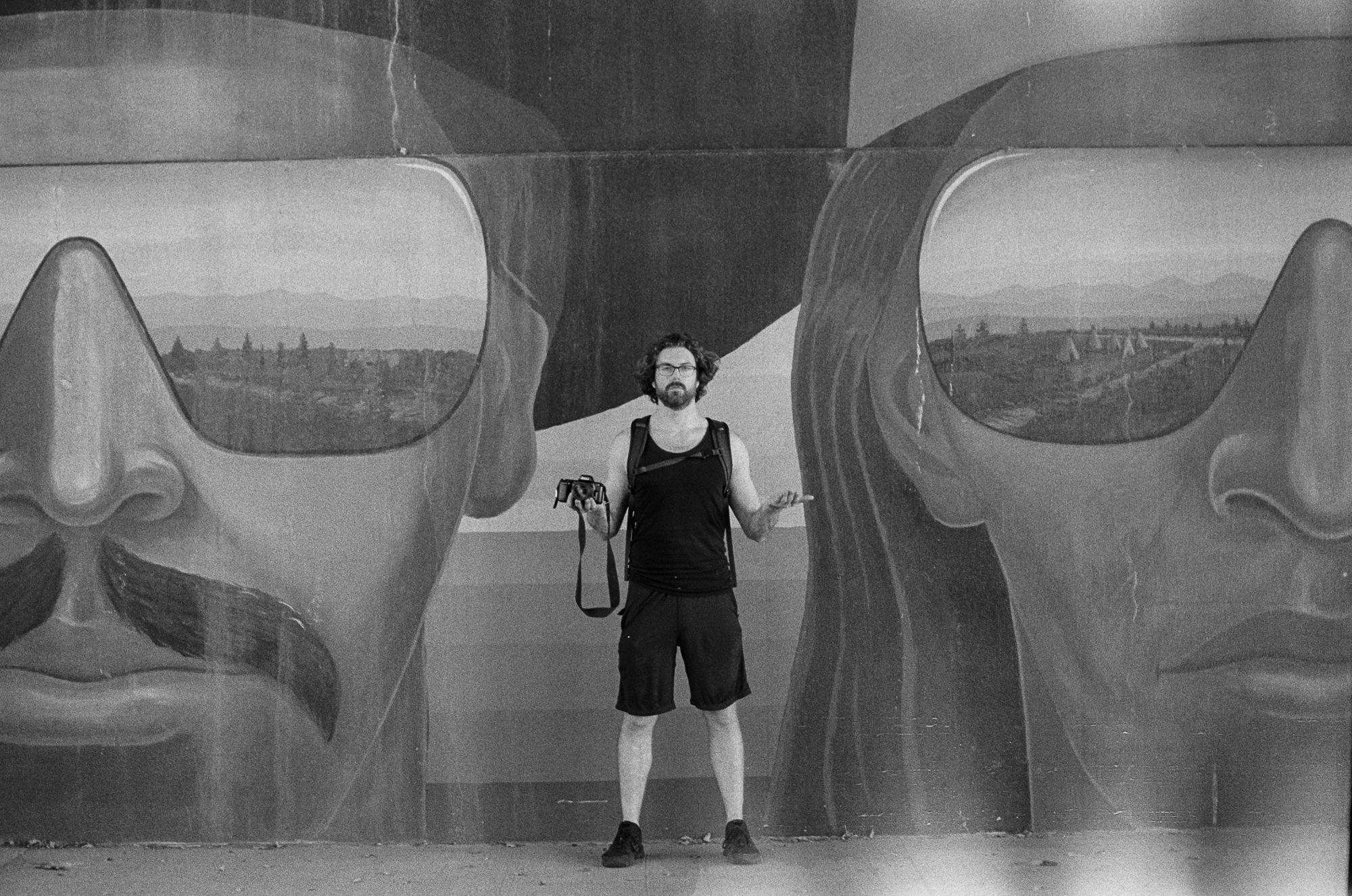 The photographer stands between two giant mural faces wearing reflective sunglasses, the Denver skyline reflected in the lenses — analog black and white 35mm film