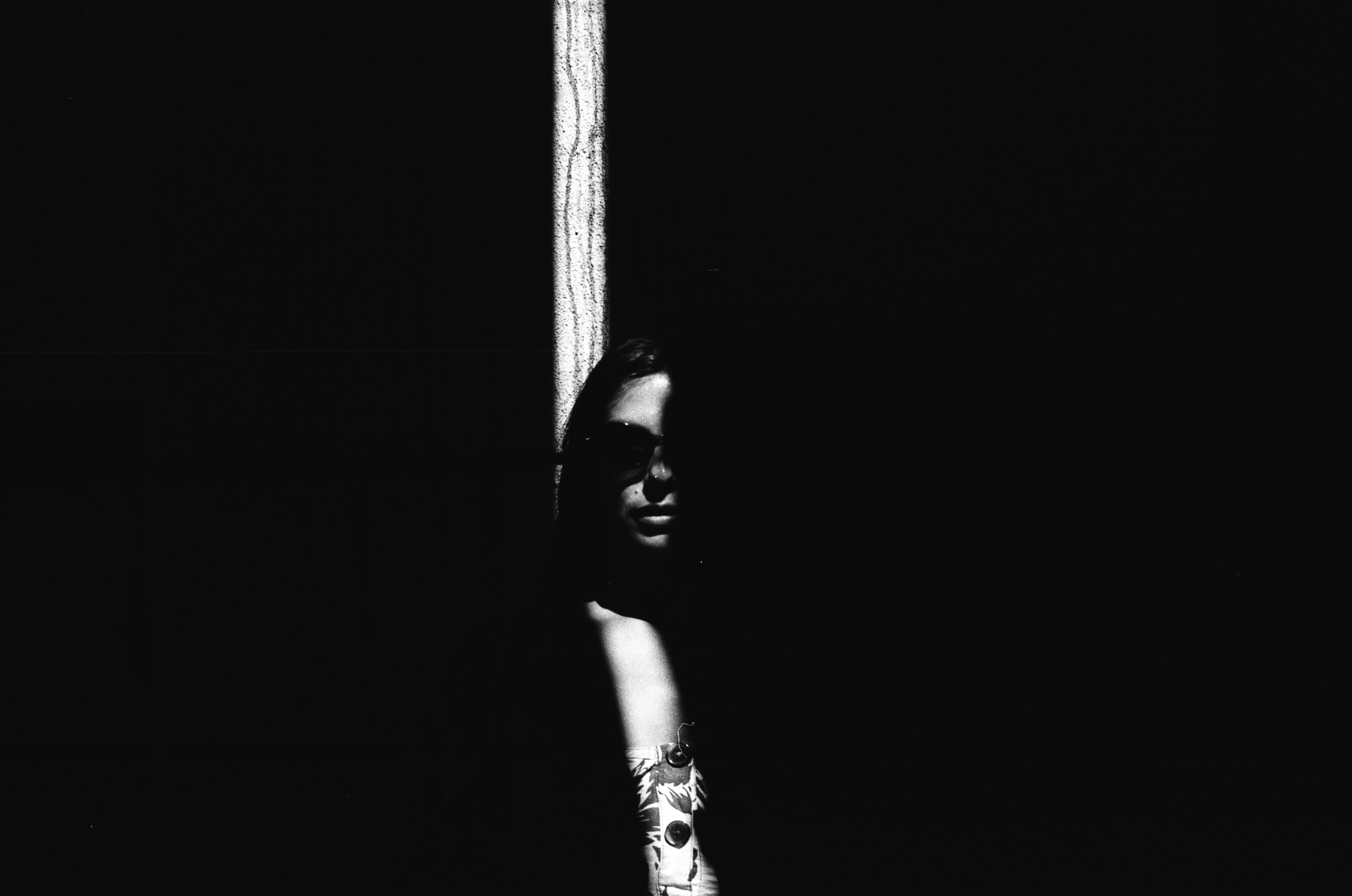 A face barely visible at the edge of near-total darkness — a single vertical strip of light on one side, extreme contrast, analog black and white 35mm film