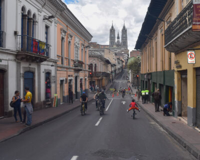 Cyclists ride down a closed street in Quito's old town, flanked by pastel colonial buildings, with the twin Gothic spires of the Basílica del Voto Nacional rising in the distance under a cloudy sky.
