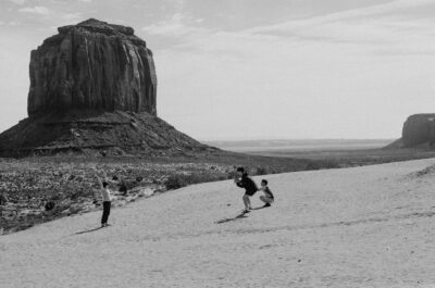 Black and white photograph of three people on a sandy slope in Monument Valley, one posing with arms raised while another crouches to take a photo, with Merrick Butte rising in the background.