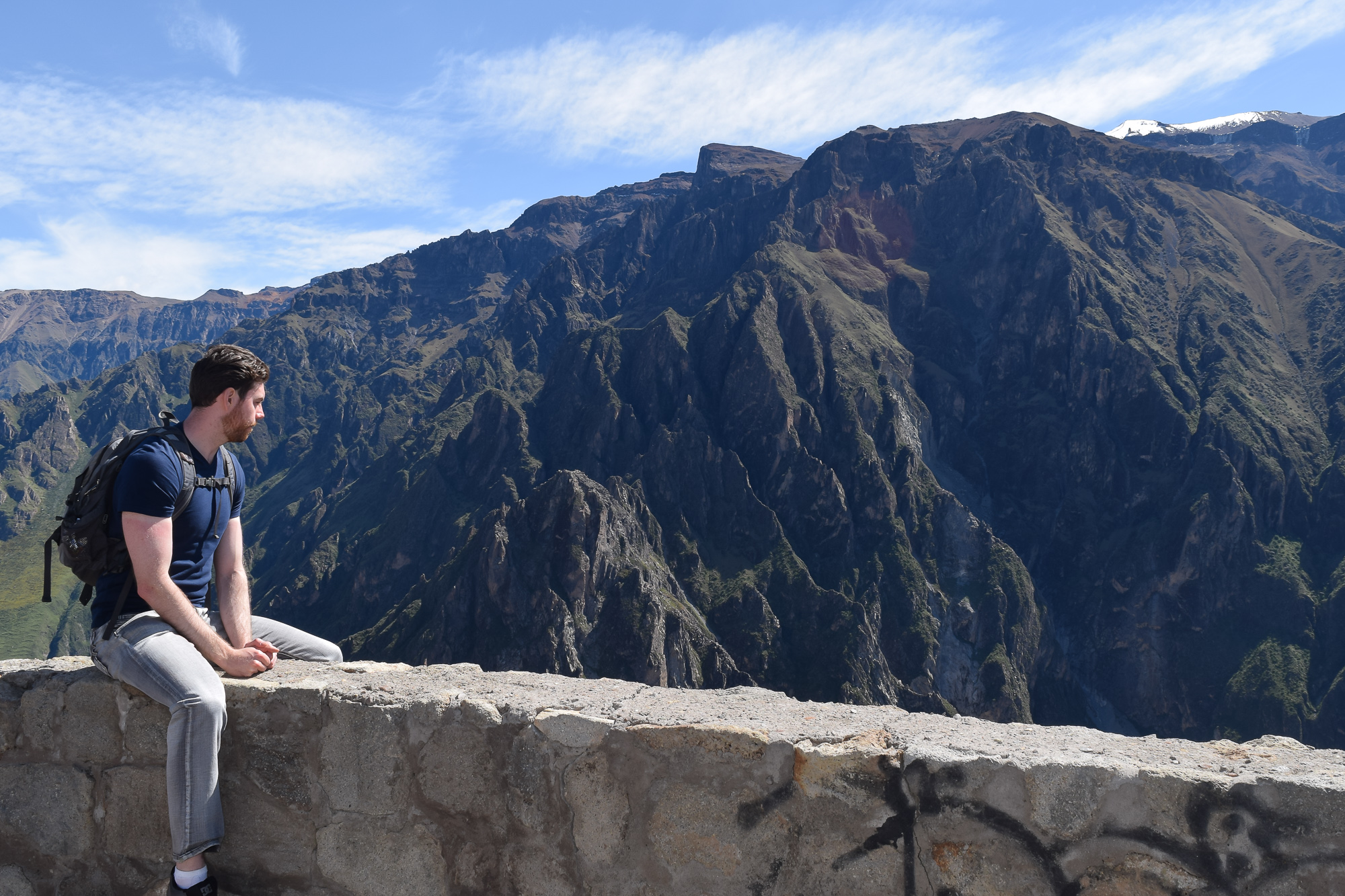 A bearded man in a navy shirt and gray jeans sits on a graffiti-marked stone wall, looking out at the rugged cliffs of a vast canyon under a blue sky.