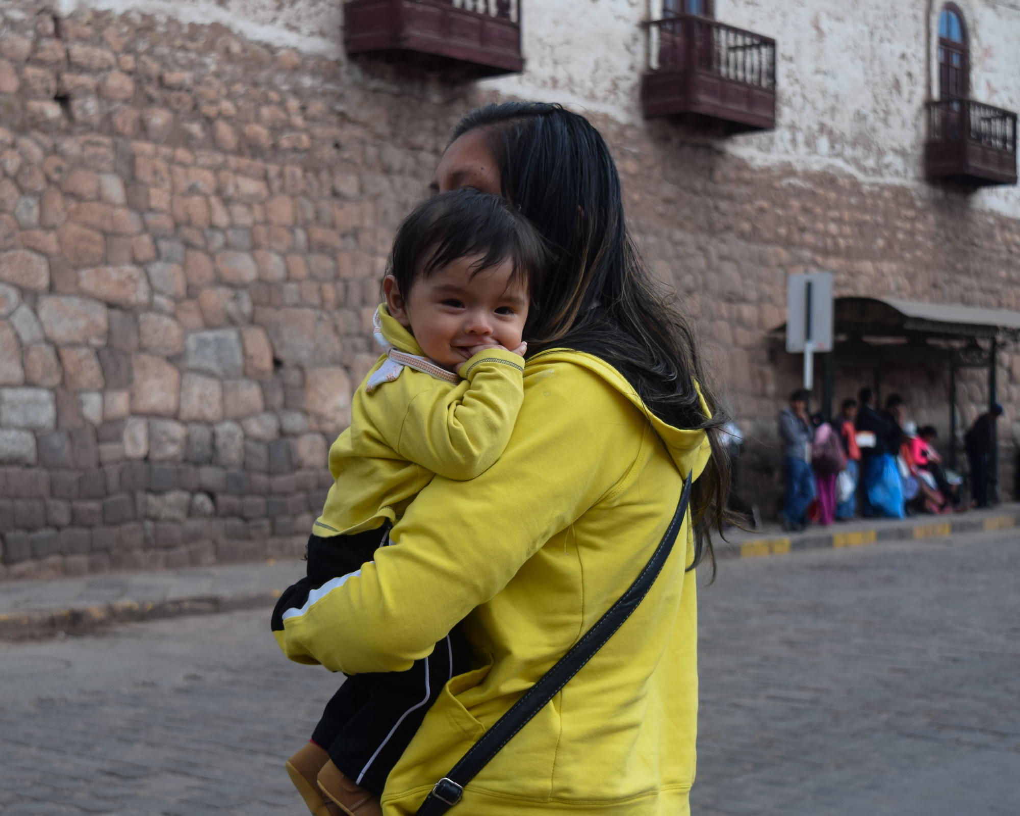 A woman in a yellow hooded sweatshirt carries a small child on her hip down a cobblestone street in Cusco. The child peeks over her shoulder, smiling with a hand near their mouth, while an Inca stone wall and colonial balconies frame the background.