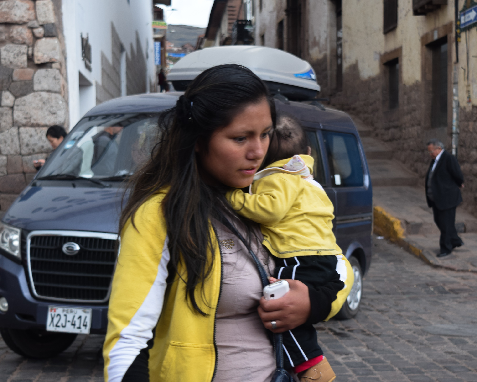 A young woman in a yellow and white jacket carries a sleeping child over her shoulder while holding a white cell phone, walking past a parked blue van on a steep cobblestone street lined with stone buildings in Peru.