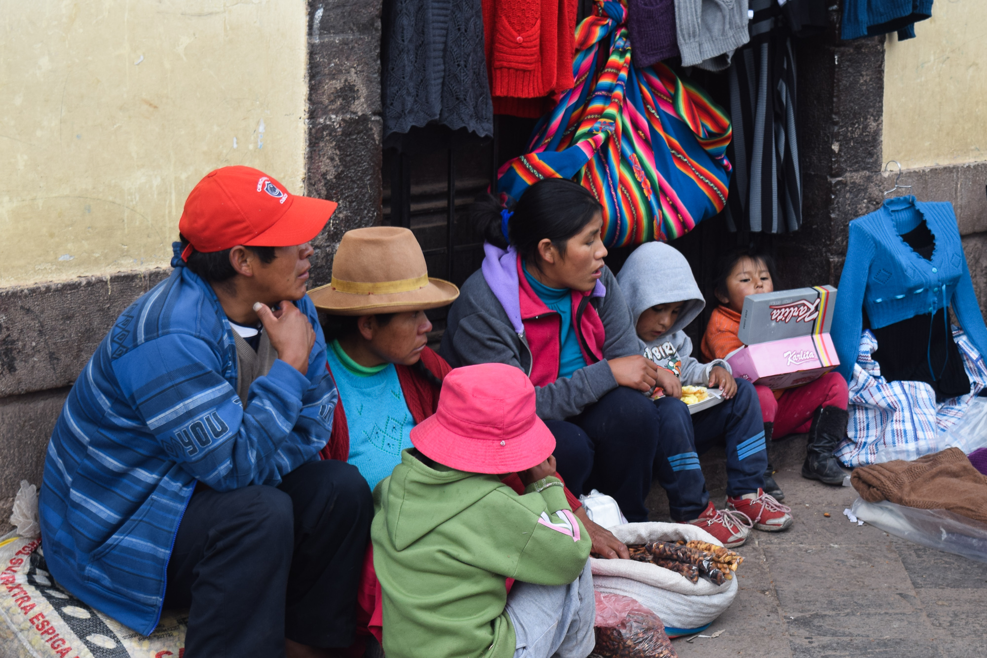 A Peruvian family sits on a stone sidewalk beside a sack of toasted corn, sharing a meal in front of a shop displaying sweaters and a striped Andean textile bag. Adults in hats and bright jackets sit alongside two small children, one holding a pink Karlita box.