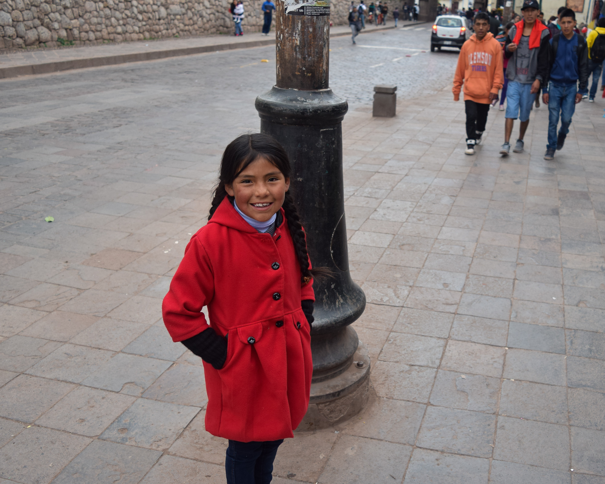 A young girl with braided hair, wearing a red coat with black buttons, stands beside a black metal lamppost on a cobblestone street in Cusco. Behind her, pedestrians walk along the sidewalk and a car drives past stone walls.