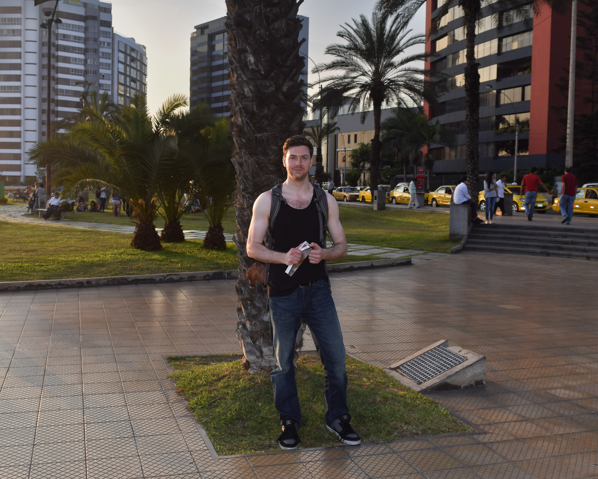 A man in a black tank top and jeans stands beside a palm tree on a tiled plaza in Lima, with high-rise buildings and yellow taxis visible in the background under warm late-afternoon light.