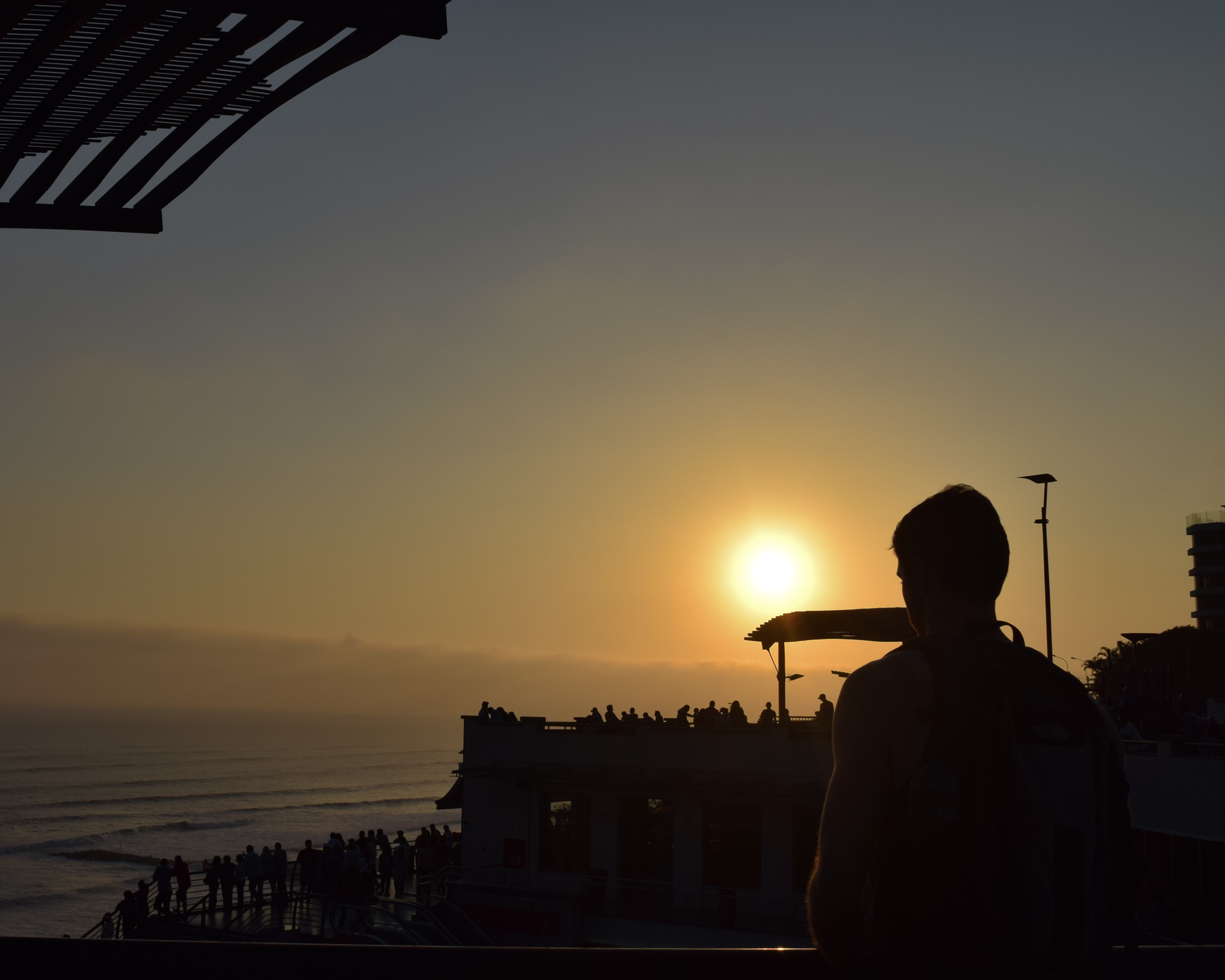 A man with a backpack stands in silhouette on a coastal terrace at sunset, looking toward the Pacific Ocean. Below him, a crowd gathers on a lower boardwalk as the sun sinks into the haze above the water.
