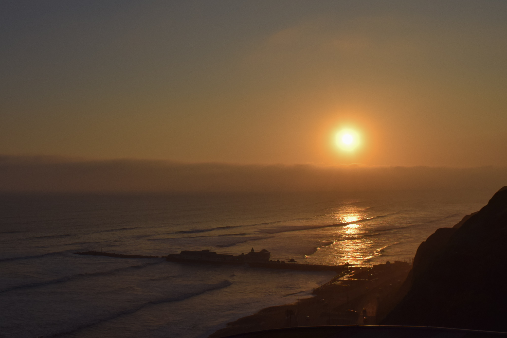 A sun low in a hazy orange sky casts a bright reflection across the Pacific Ocean, with a rocky cliff silhouetted on the right and a small coastal settlement visible along the shoreline below.
