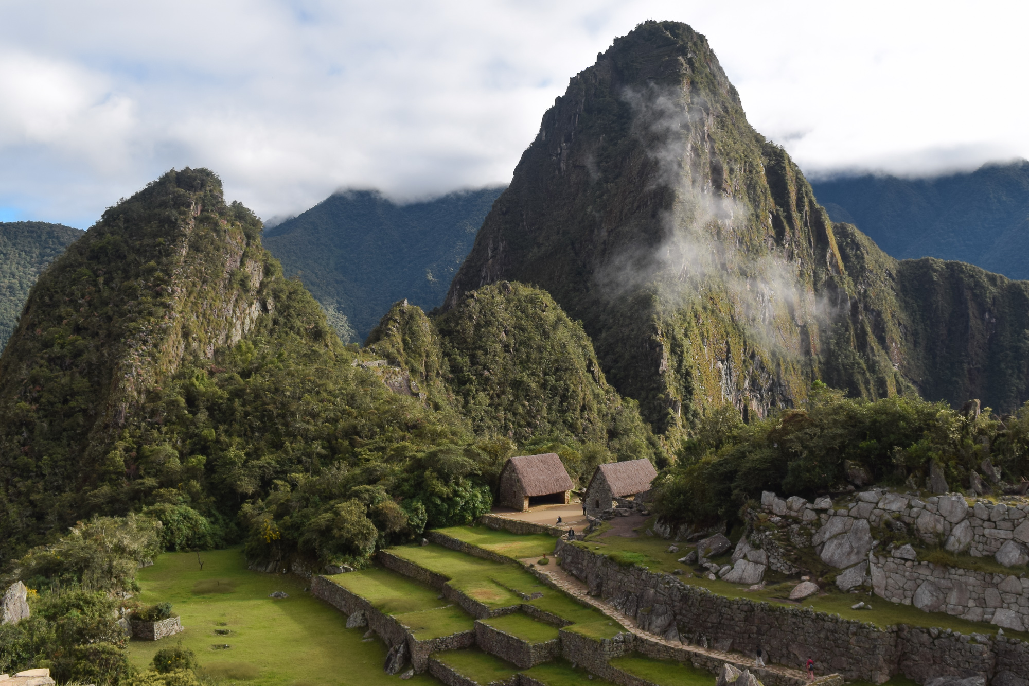Inca stone terraces and two thatched-roof stone buildings sit on a green plateau below the steep, mist-wrapped peak of Huayna Picchu in Peru.