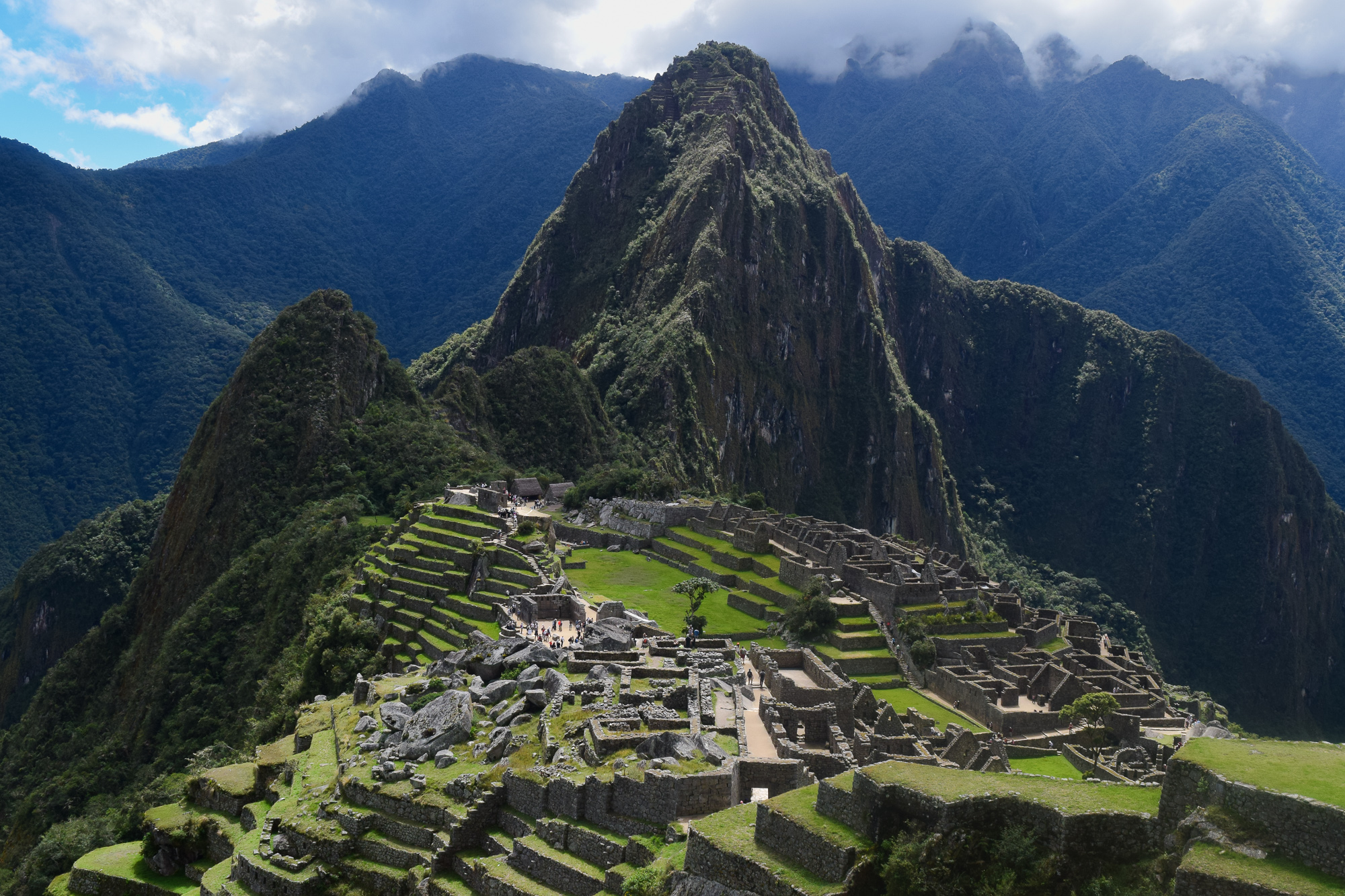 Stone terraces and ruins of Machu Picchu spread across a ridge, with the steep green peak of Huayna Picchu rising behind under a partly cloudy sky.
