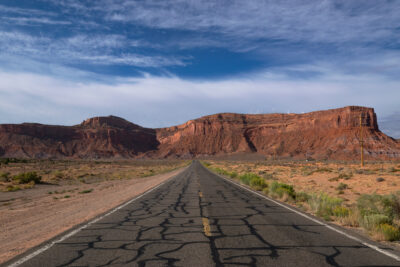A weathered two-lane asphalt road with heavy crack repairs runs straight toward distant red sandstone cliffs under a blue sky streaked with cirrus clouds. Sparse desert scrub and a line of wooden utility poles flank the highway.