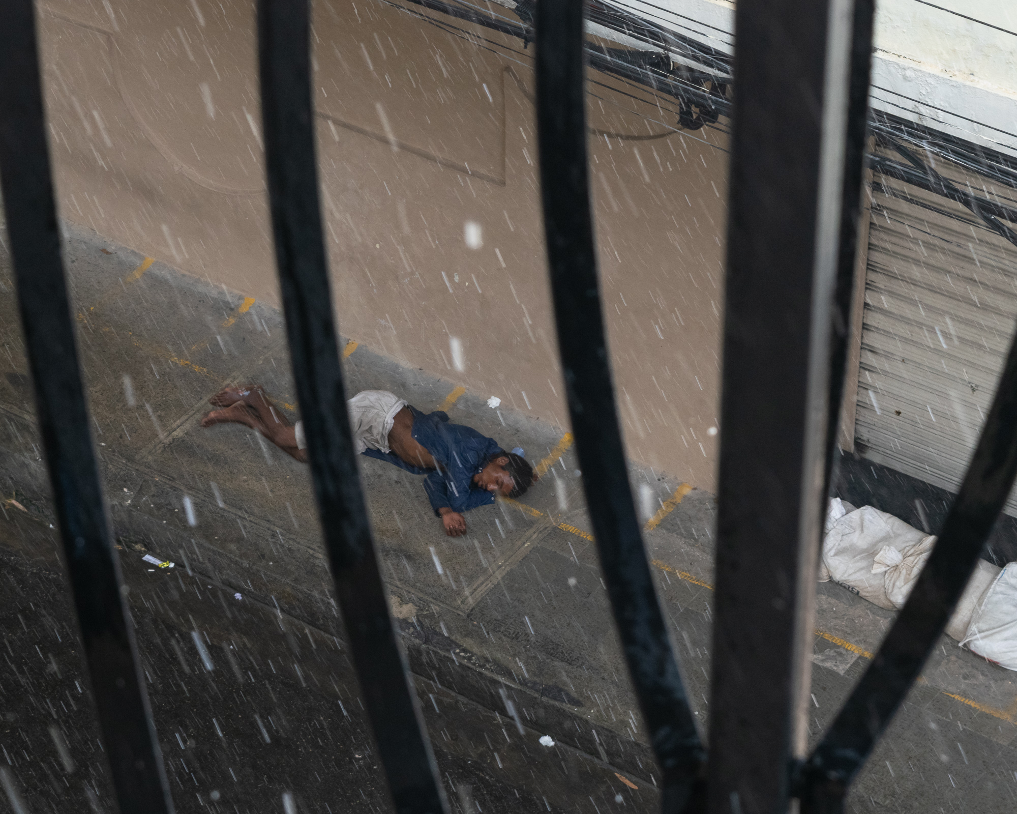 Viewed through the vertical bars of a wrought iron gate, a man in a blue jacket and light pants lies curled on a wet sidewalk as rain falls. A shuttered storefront and white sacks of refuse are visible beside him.