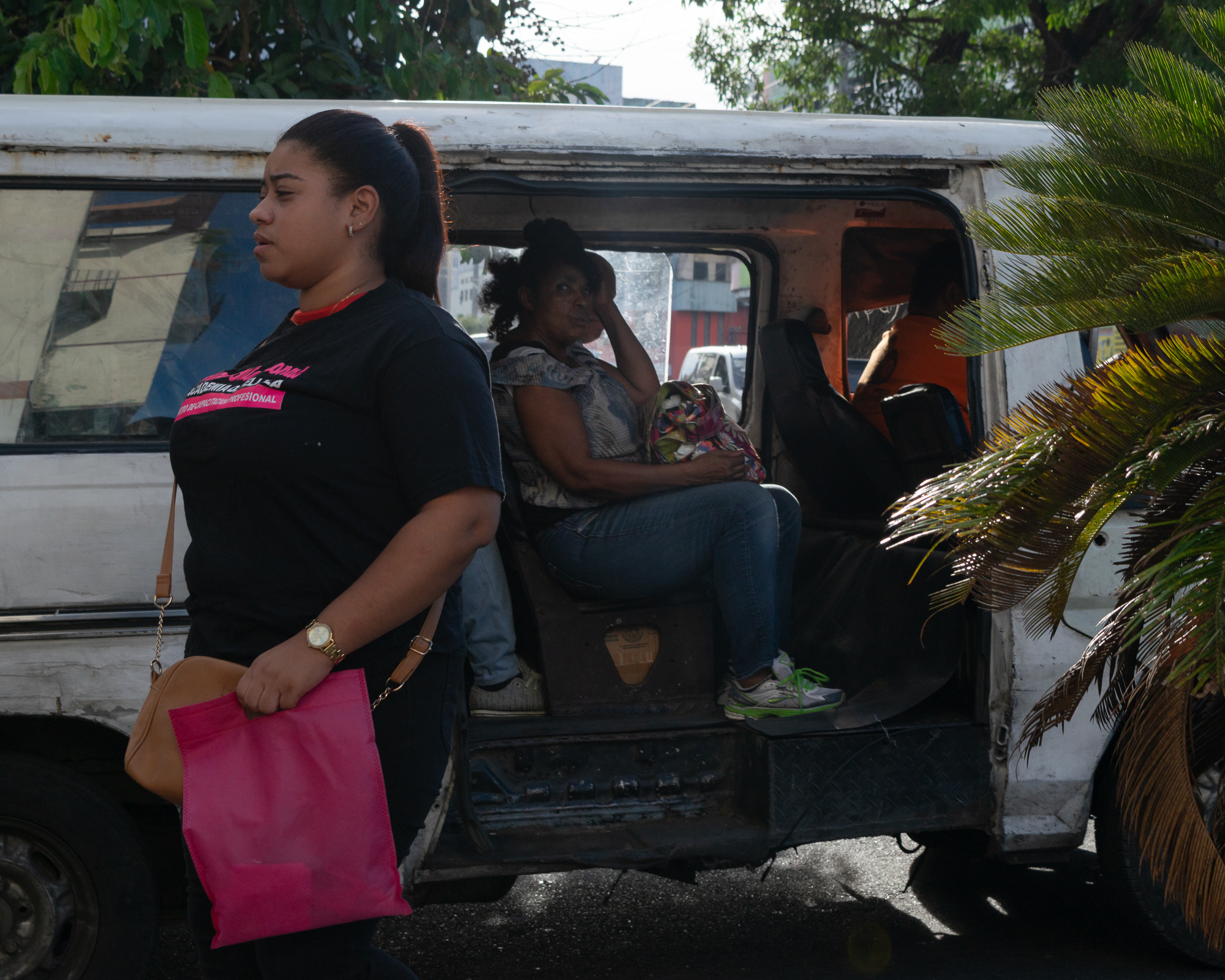 A young woman in a black t-shirt carries a bright pink bag past the open side door of a battered white passenger van, where an older woman sits waiting with a floral purse on her lap. Morning light filters through palm fronds on the right side of the frame.