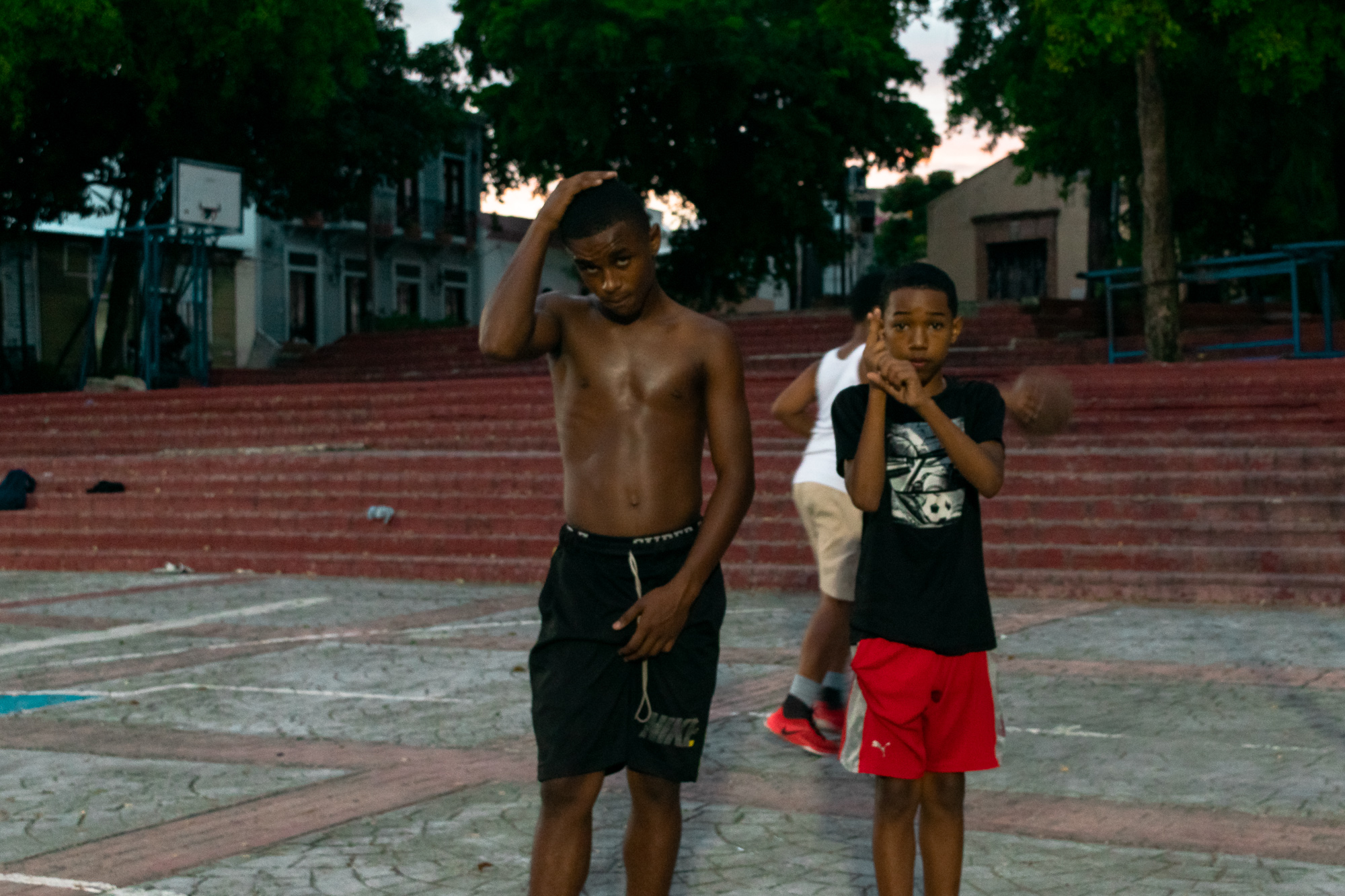 A shirtless teenage boy in black Nike shorts stands on a tiled plaza beside a younger boy in a black t-shirt and red Puma shorts, while a third kid in a white tank top shoots a basketball near the red concrete steps behind them.