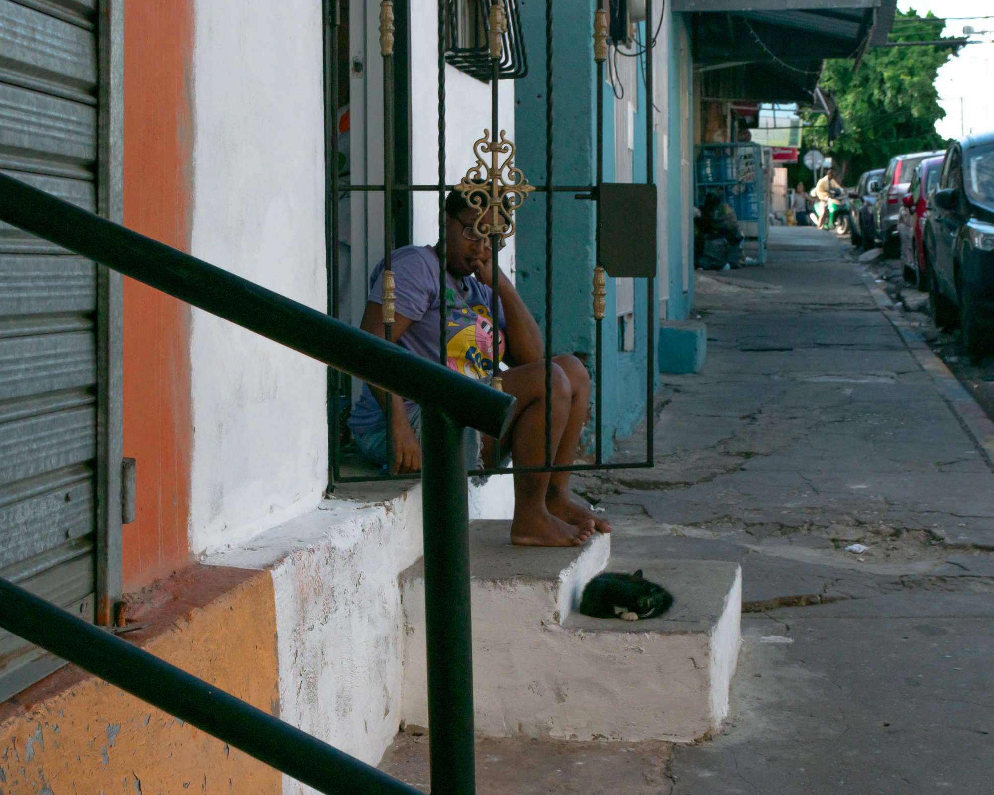 A barefoot woman in a purple t-shirt sits behind a wrought iron gate on a white concrete stoop, resting her chin on her hand, while a black and white cat sleeps curled on the same step. The sidewalk stretches past colorful storefronts toward parked cars and a distant motorcyclist.