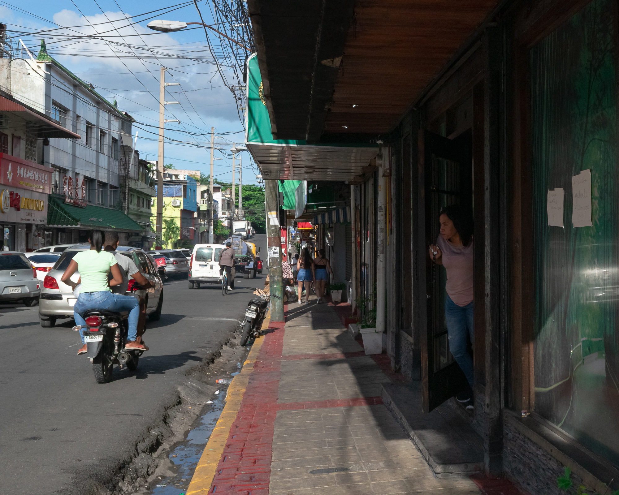 A woman in a pink shirt and jeans leans in a shop doorway watching traffic on a busy Santo Domingo street, while two riders share a motorbike and tangled power lines crisscross the sky above.