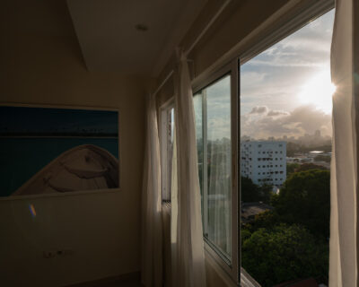 A framed photograph of a wooden boat on turquoise water hangs on a beige wall beside a sliding glass window. Through the window, late afternoon sun breaks over the Santo Domingo skyline, a white apartment block, and dense treetops.