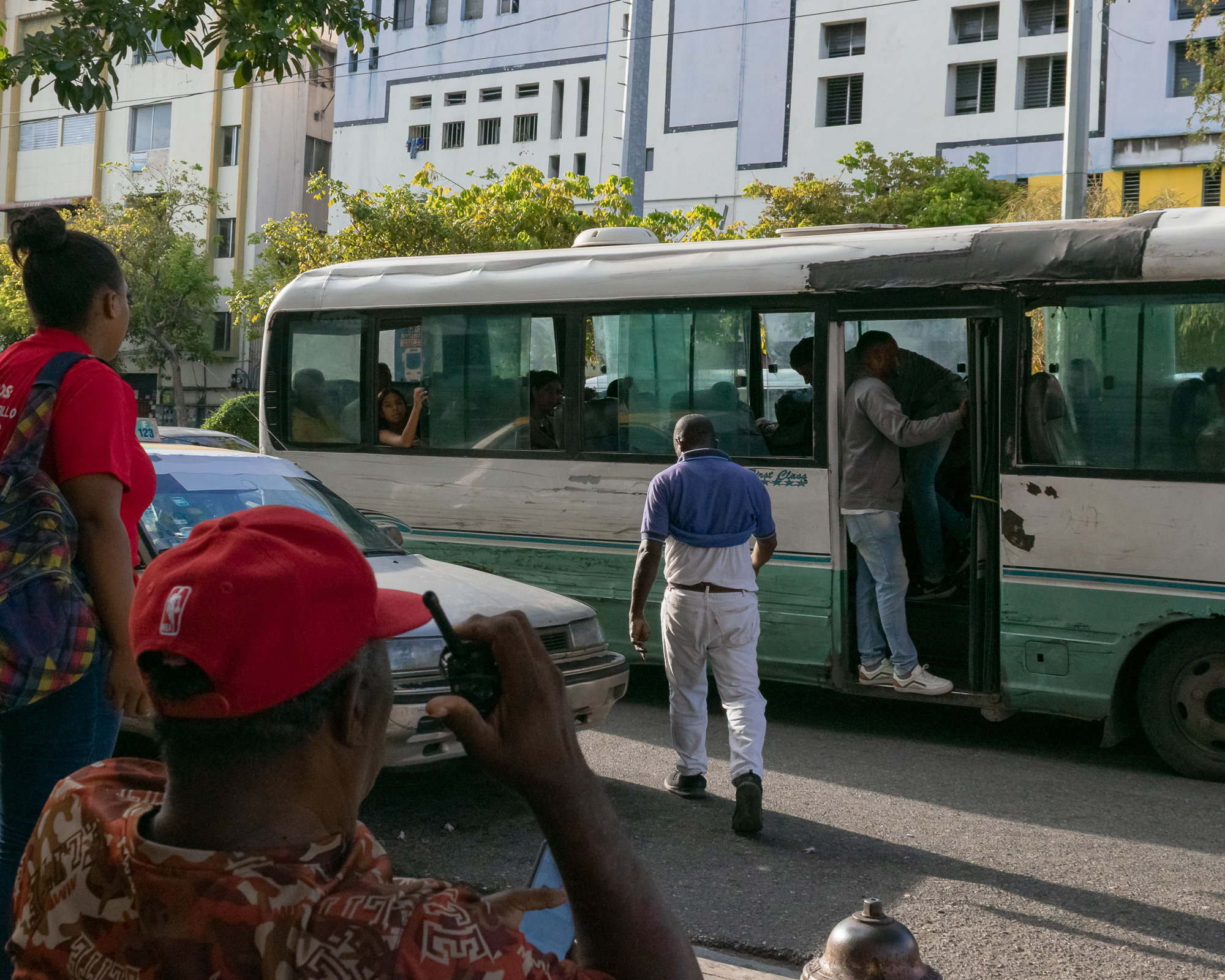 A man in a blue polo and white pants walks toward a green and white guagua bus where a young man hangs from the open doorway. In the foreground, a man in a red NBA cap holds a walkie-talkie while a woman in a red shirt watches the bus pass.