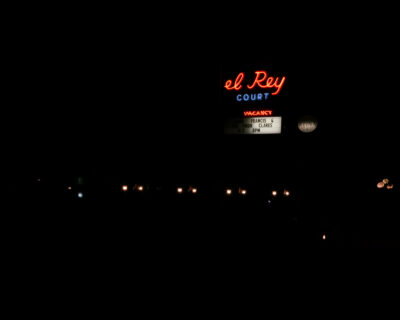 A neon sign reading 'el Rey Court' glows red and blue against a black night sky, with a marquee below announcing John Francis and the Poor Clares on 9/7 at 8PM. Small lamp lights line the ground below the sign.