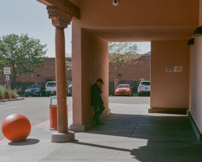 A man in a dark jacket and cap leans against an adobe column under a covered walkway, smoking. An orange concrete bollard sits in the foreground, with parked cars visible through the archway.