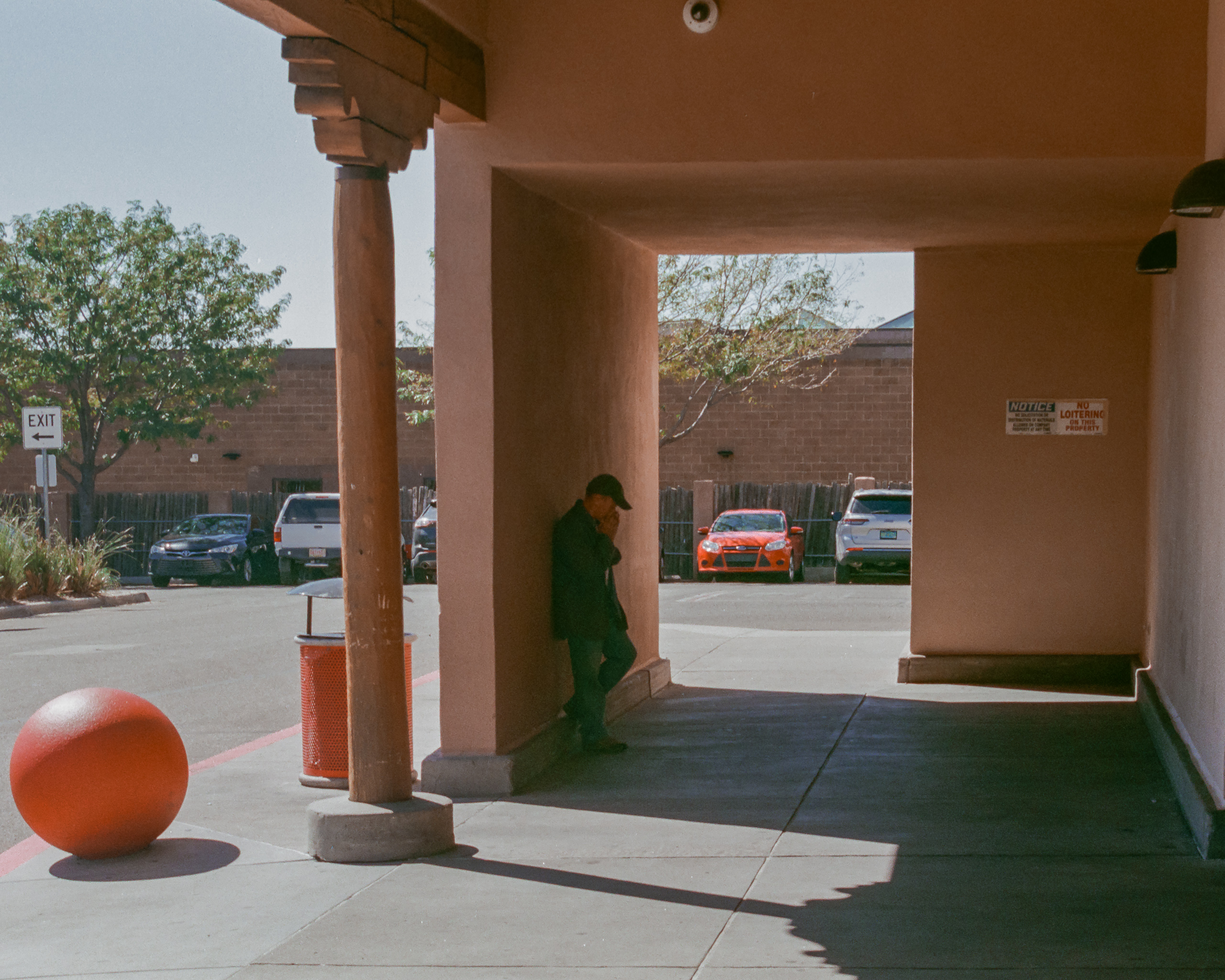 A figure stands in the shade of an adobe portico in Santa Fe, New Mexico, as hard diagonal shadows cut across the concrete below