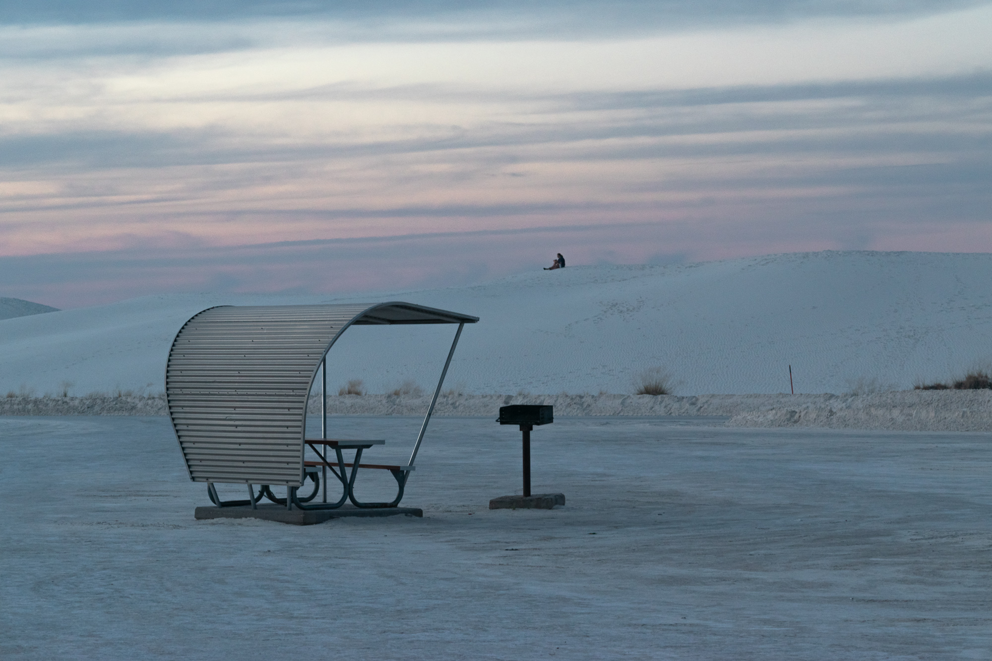 White Sands New Mexico at dusk — a metal picnic shelter and grill on the gypsum flat, a lone figure seated on a distant dune beneath a pink and blue sunset sky
