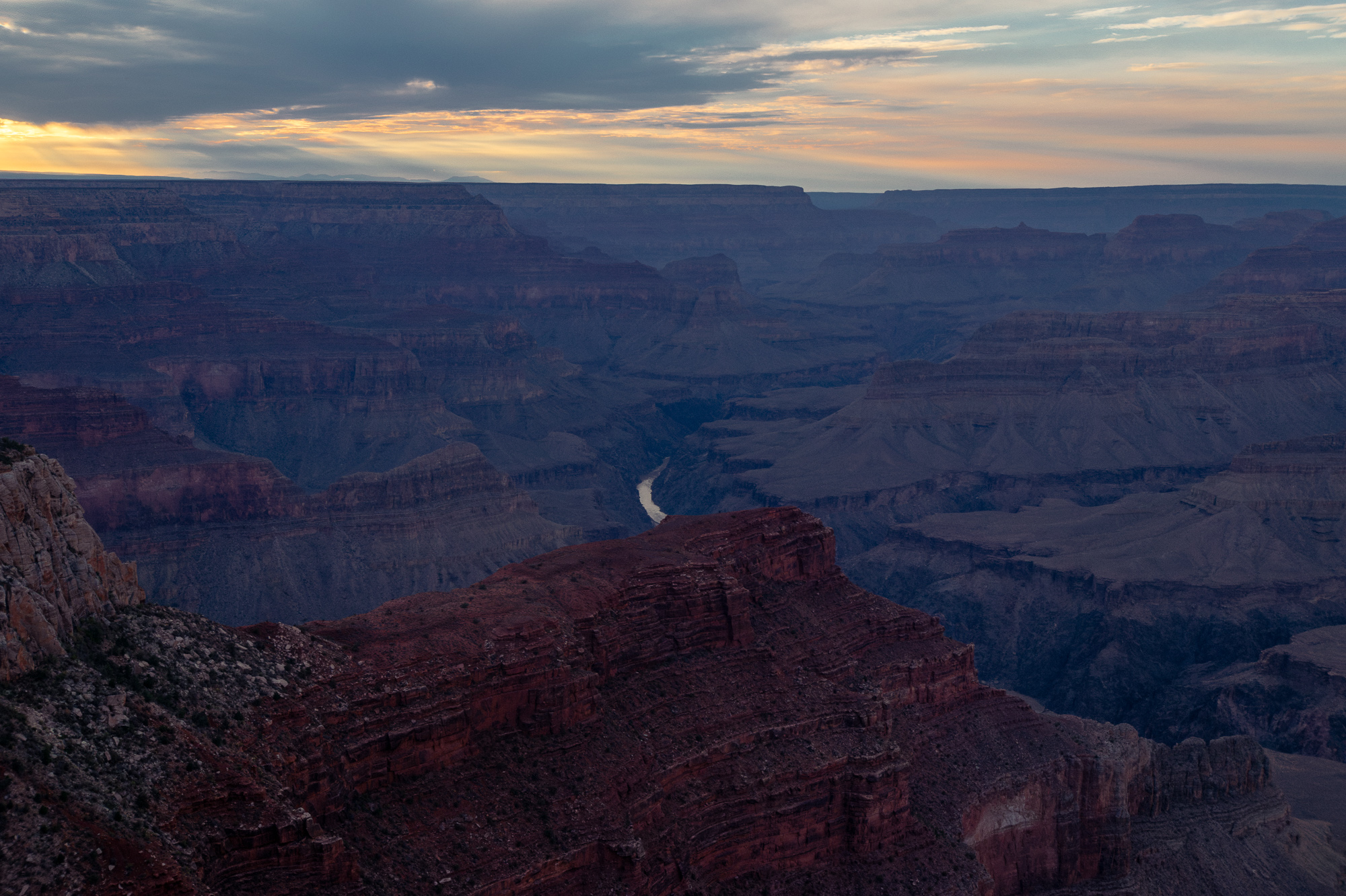 A wide dusk view into the Grand Canyon, with layered blue-grey buttes receding to a flat horizon under a streaked orange and grey sky. A pale ribbon of the Colorado River is visible far below in the shadowed gorge, while a red sandstone ridge fills the foreground.
