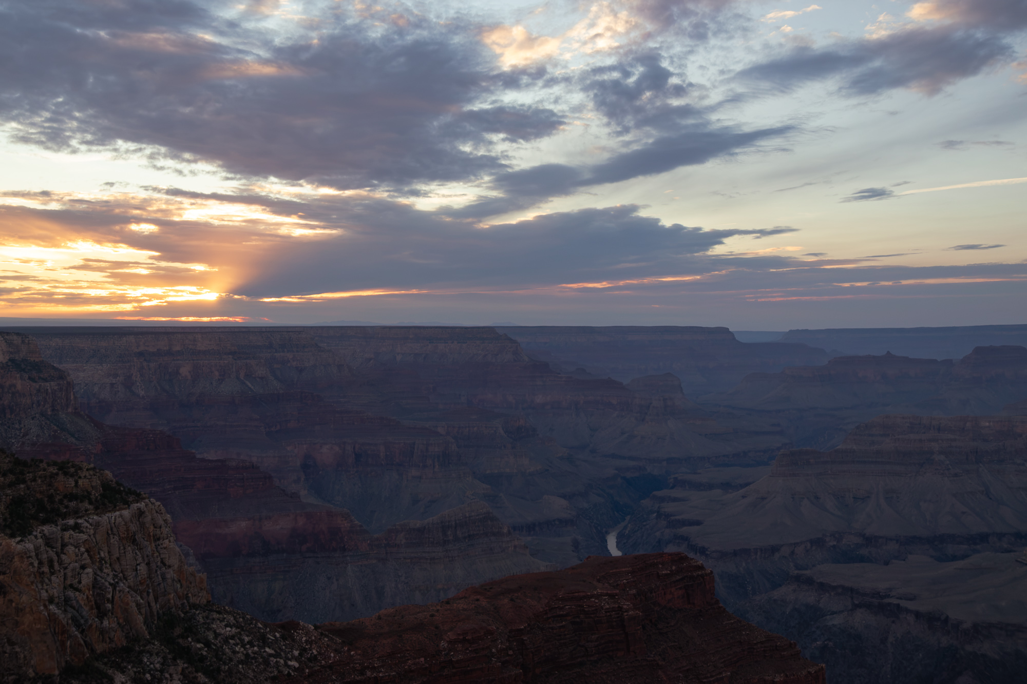 A wide view across the Grand Canyon at sunset, with layered red and gray buttes receding into haze and a thin bend of the Colorado River visible in the shadowed depths. Above the flat horizon, a break in the cloud deck lets orange light fan out across a sky of gray and lavender cloud bands.