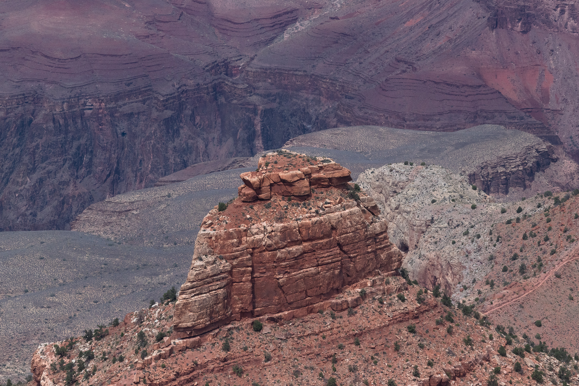A blocky red sandstone butte rises from a scrub-dotted ridge in the Grand Canyon, its horizontal rock layers catching flat afternoon light. Behind it, pale gray talus slopes and hazy purple-toned cliff terraces recede into the canyon depths.