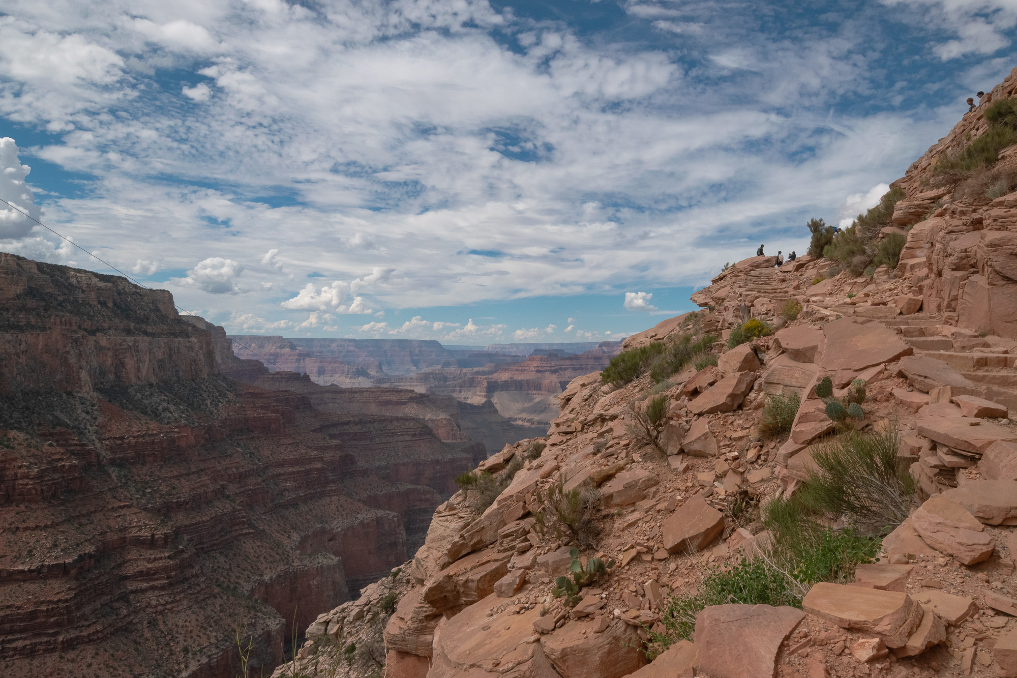 A wide view from a rocky trail cut into red sandstone on the right, where several small figures sit and stand on a ledge near scattered shrubs and prickly pear. To the left the Grand Canyon drops away in layered cliffs and terraces receding into blue haze, under a sky filled with broken white cloud.