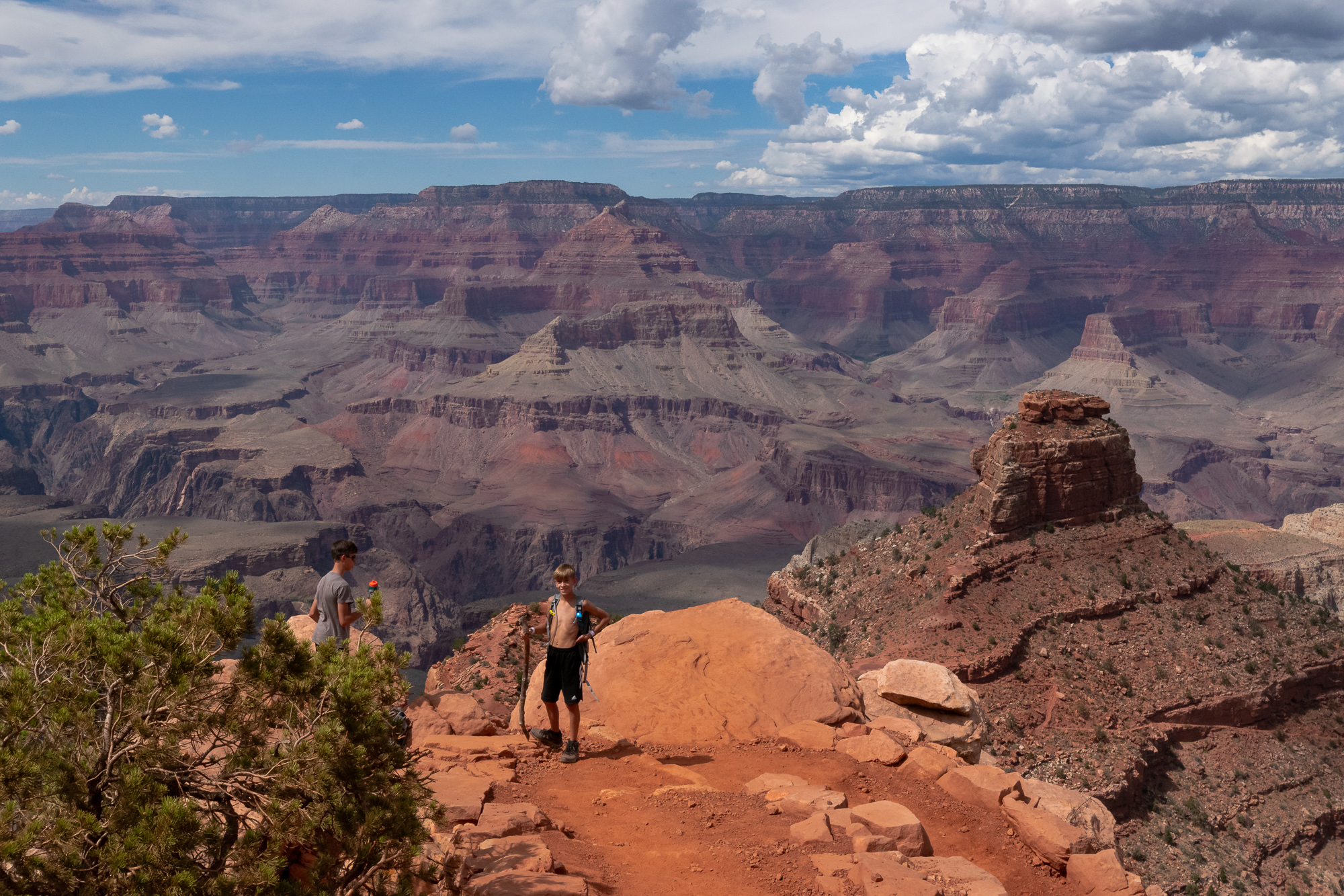 A shirtless boy with a hiking stick and small backpack stands on a rust-colored sandstone outcrop above the Grand Canyon, hand on hip, while an older boy in a grey t-shirt looks out over the rim to the left. Behind them, layered red and grey buttes recede toward a flat plateau under tall cumulus clouds.