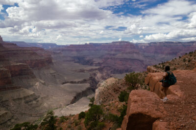 A woman in a dark tank top and shorts with a teal backpack and a camera stands on red sandstone boulders at the right edge of a canyon rim, looking out over layered rock terraces that drop into a wide gorge. Cumulus clouds fill most of the sky above the distant plateau walls.