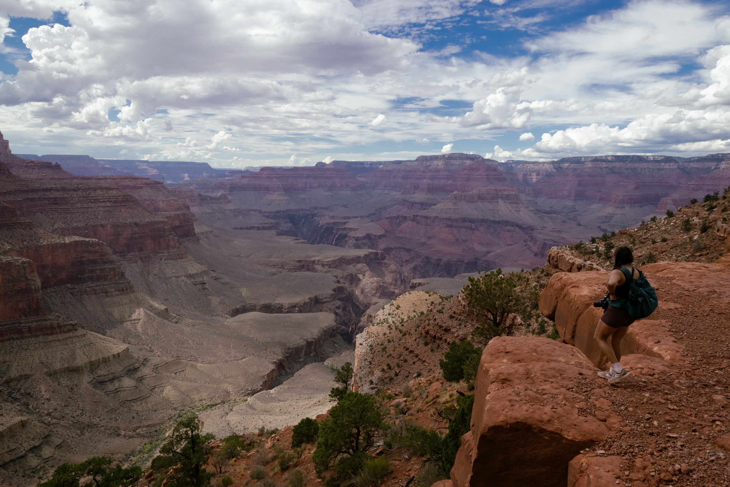 A woman in a dark tank top and shorts with a teal backpack and a camera stands on red sandstone boulders at the right edge of a canyon rim, looking out over layered rock terraces that drop into a wide gorge. Cumulus clouds fill most of the sky above the distant plateau walls.