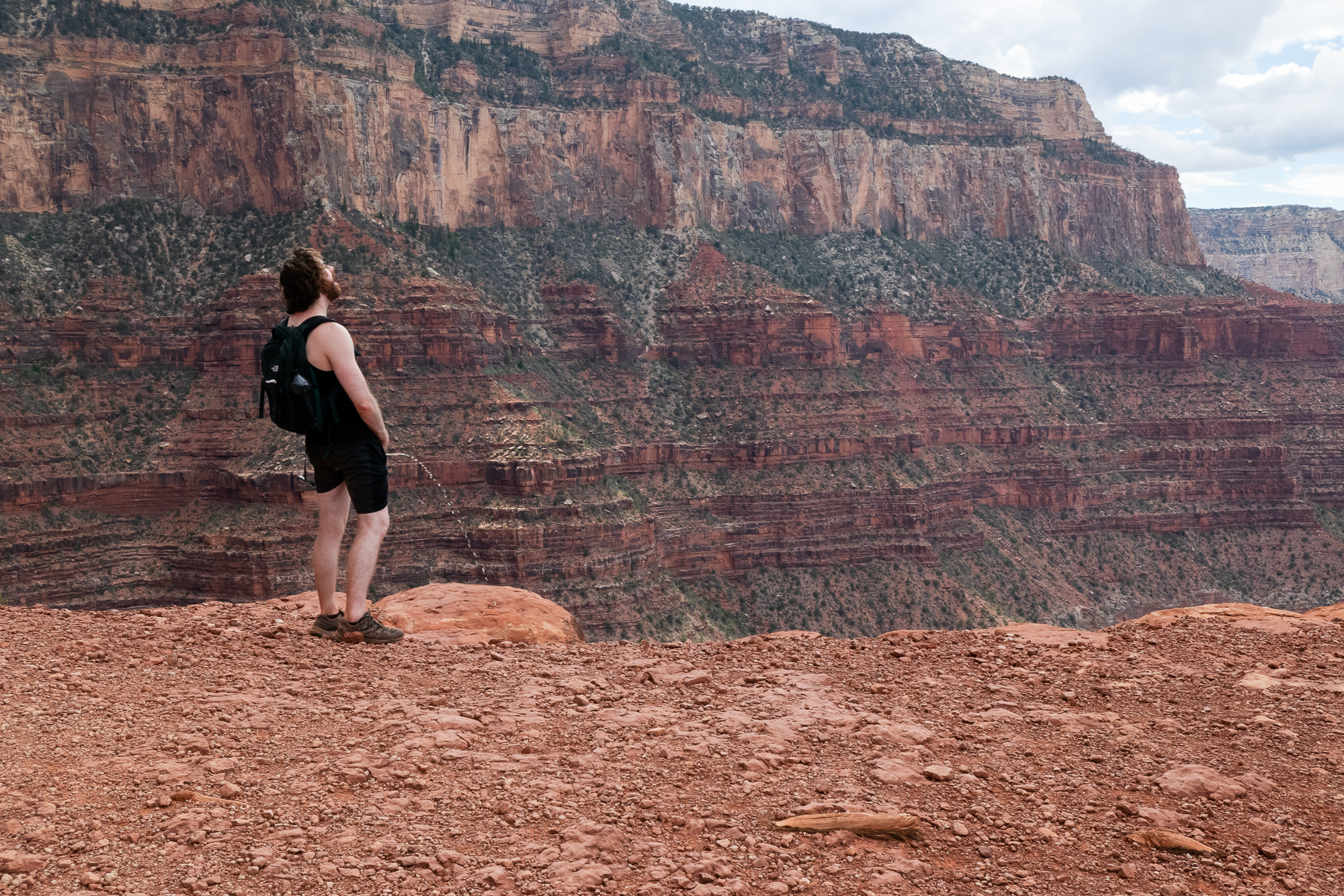 A bearded man in a black tank top, black shorts and a dark backpack stands on red sandstone at the edge of the Grand Canyon, facing away from the camera with a thin arc of liquid falling into the void. Behind him, banded red and tan cliff walls rise in layers toward a cloudy sky.