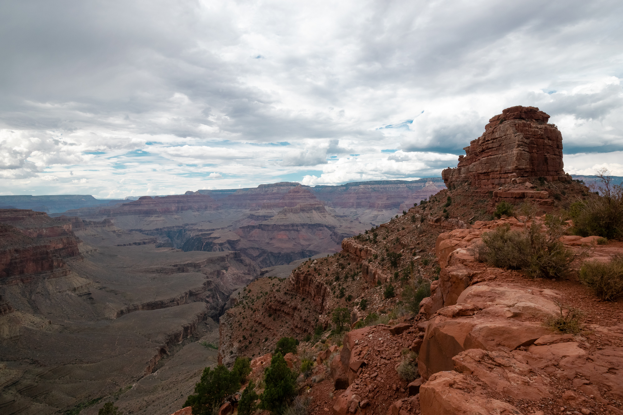 A wide view of the Grand Canyon under a heavy overcast sky, with a blocky red sandstone butte rising on the right above a rubble-strewn ridge dotted with low shrubs and juniper. To the left, layered gray and mauve canyon walls recede toward flat-topped mesas on the far horizon.