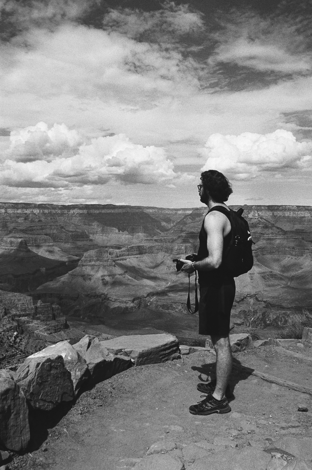 A bearded man in a dark tank top, shorts and trail shoes stands in profile on a flat rock ledge at the Grand Canyon, a camera and strap held in his hands and a daypack with a water bottle on his back. Behind him the canyon drops away in layered terraces toward a flat horizon, with large cumulus clouds filling the upper half of the black and white frame.